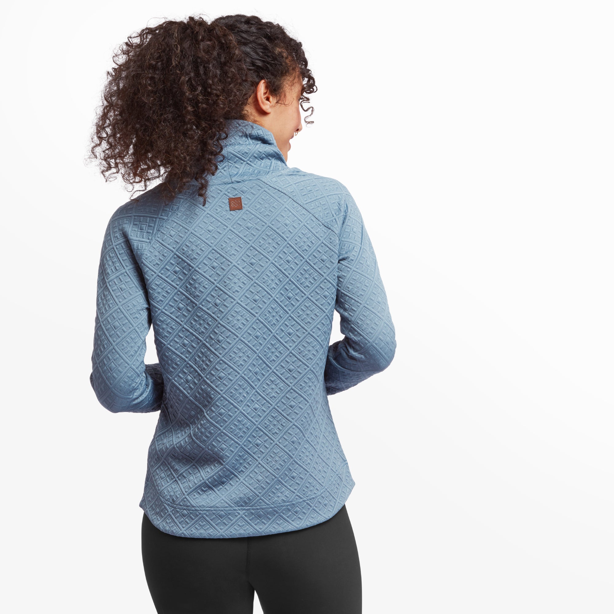 A view from the back of the Sherpa Adventure Gear Nyano Pullover in Blue, displaying the quilted diamond pattern and the detailed stitching. The small leather logo patch is visible at the top center of the back. Her curly hair is tied back, allowing the collar and pattern to stand out.