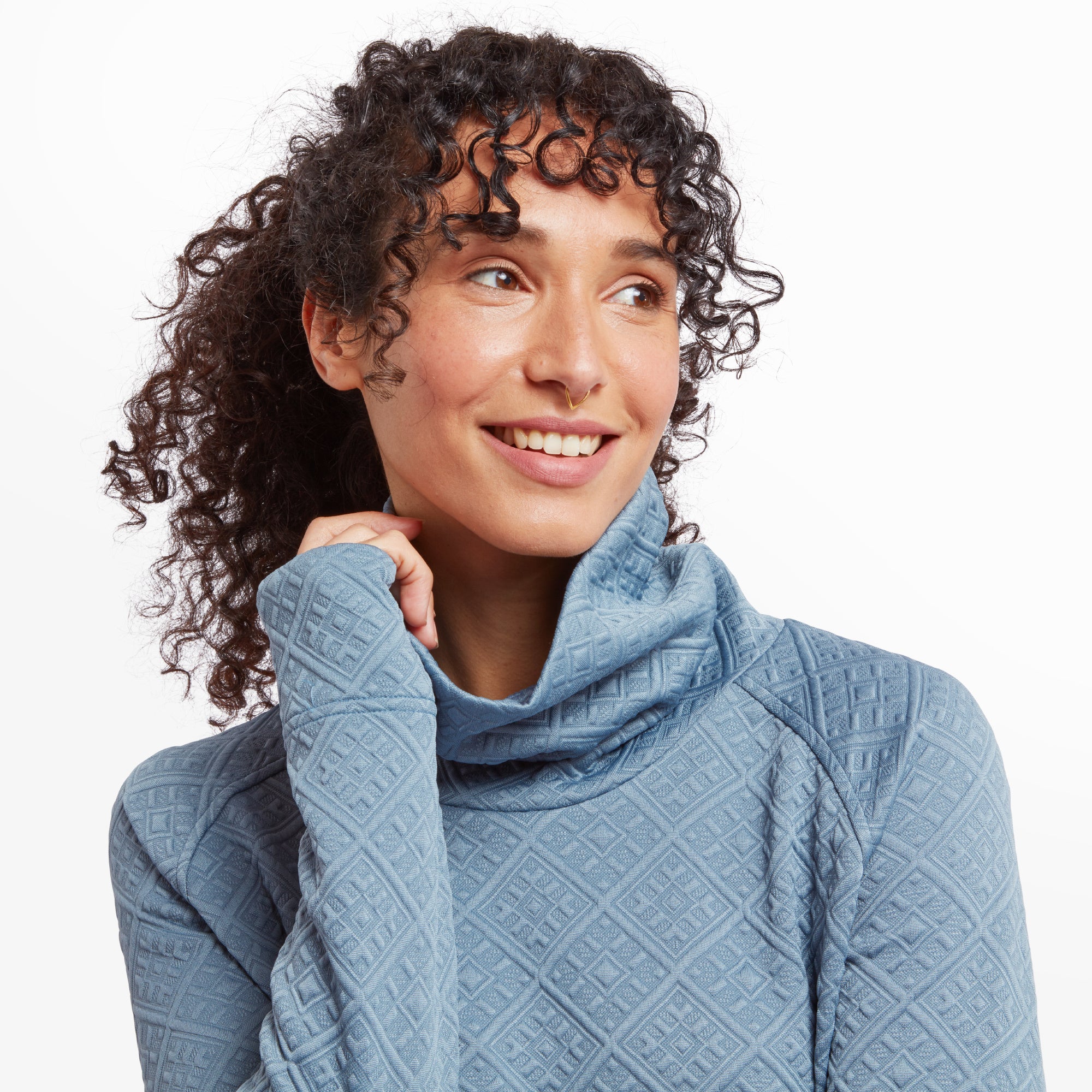 A close-up shot highlights the cowl neck design and the soft, textured fabric. The model smiles warmly, resting one hand lightly near her chin, emphasising the Sherpa Adventure Gear Nyano Pullover in Blue's warmth and comfort.