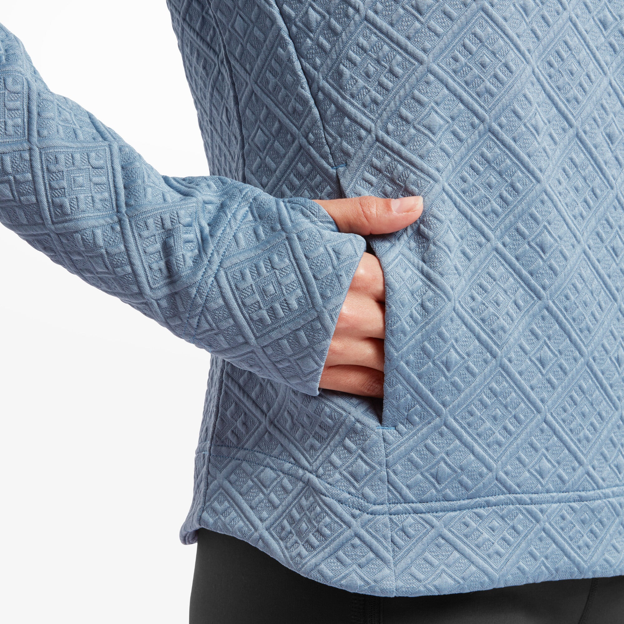 A detailed shot focuses on the side pocket of the Sherpa Adventure Gear Nyano Pullover in Blue. The model's hand is partially tucked into the pocket, showcasing the clean stitching and seamless integration of the pocket into the quilted design. The fabric's diamond pattern is vividly displayed.