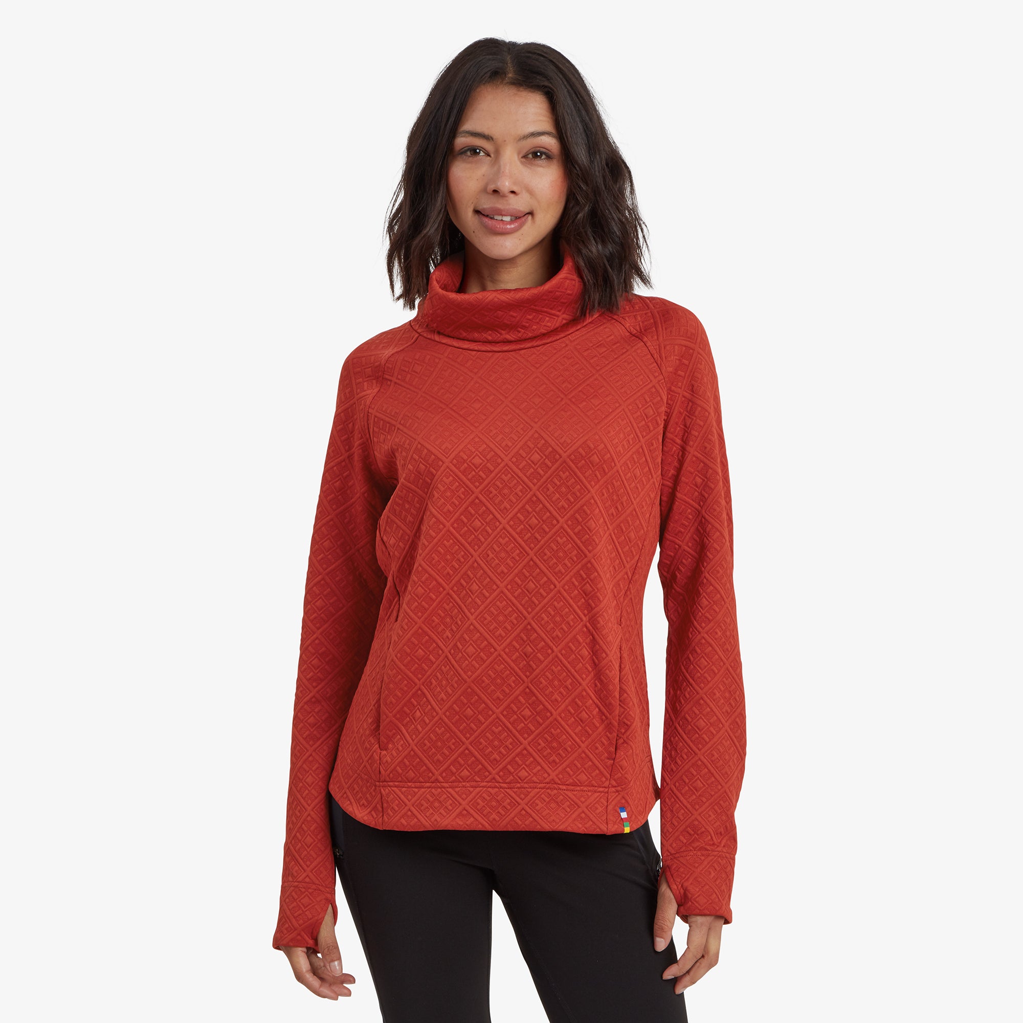 A woman wearing a Sherpa Adventure Gear Nyano Pullover in Red with a relaxed cowl neck. The textured fabric features a diamond pattern, and she pairs the top with black leggings, standing confidently with a warm smile.