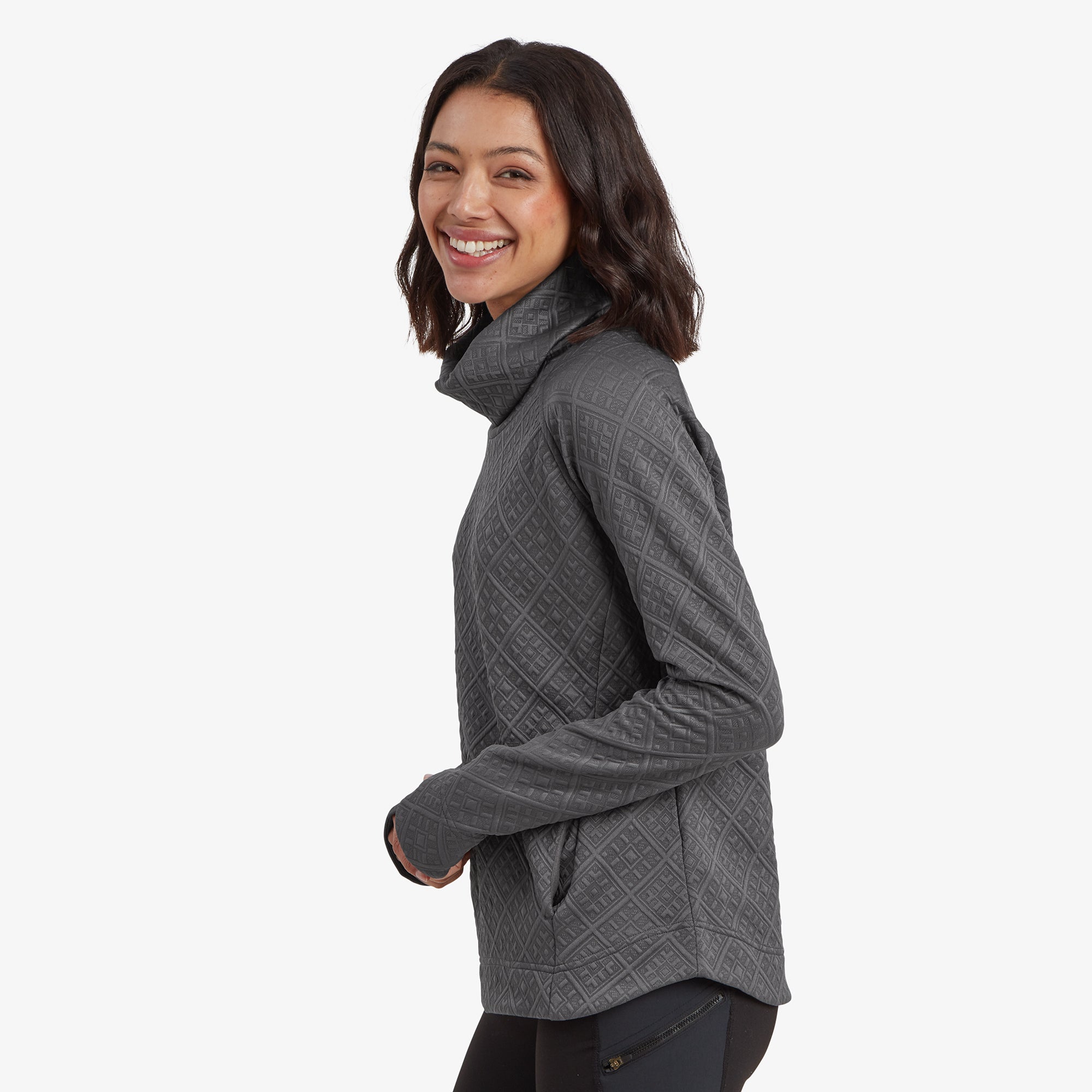 A side view of the model wearing the Sherpa Adventure Gear Nyano Pullover in Grey, standing in a relaxed pose with one hand in her pocket. The diamond-patterned texture is visible across the sleeve and body, and the draped cowl neck falls gently around her neck. She smiles while looking back over her shoulder.