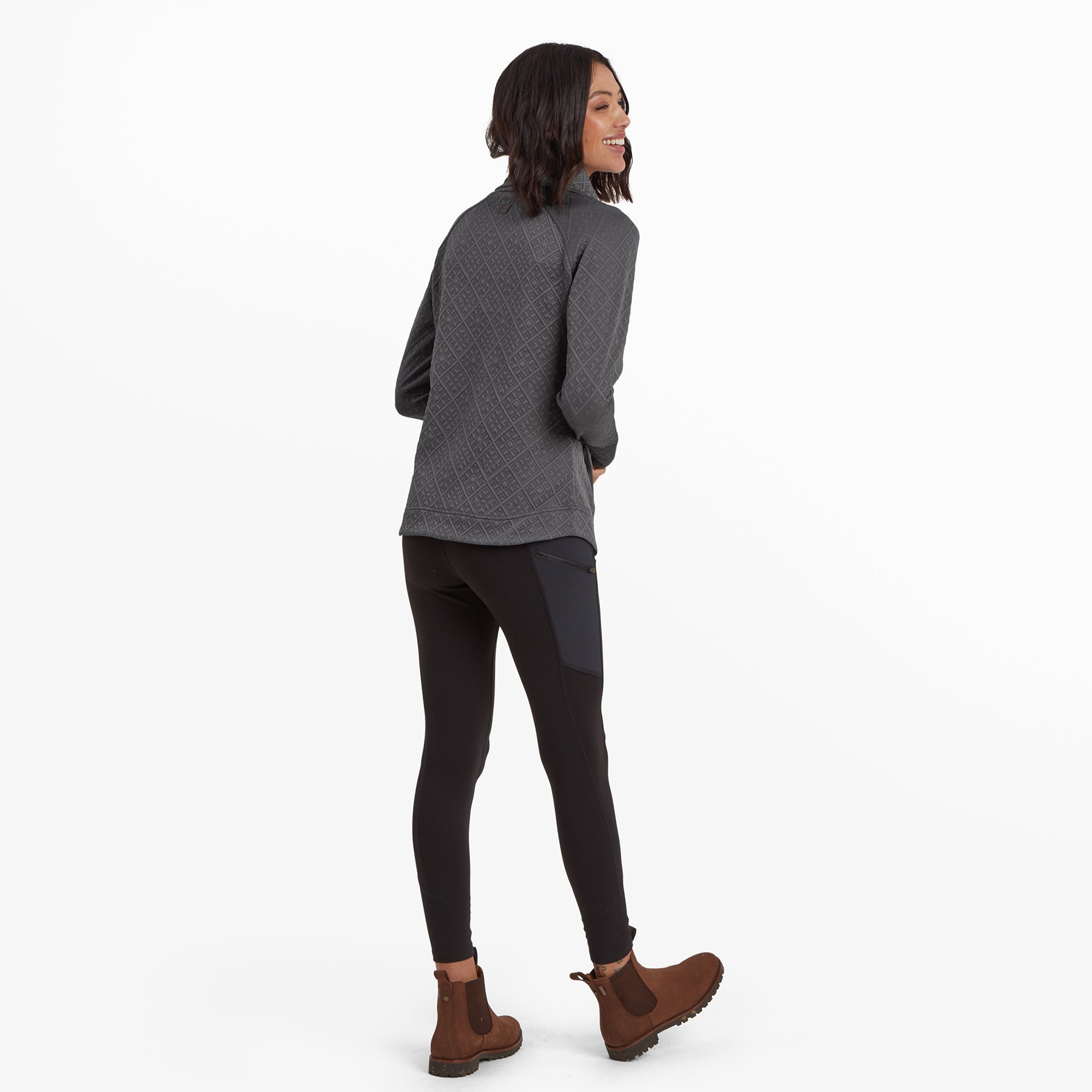 The model faces away from the camera, showcasing the back of the Sherpa Adventure Gear Nyano Pullover in Grey. The diamond pattern continues across the back, and the pullover drapes comfortably over her hips. She pairs the pullover with black leggings and brown boots with blue elastic panels.