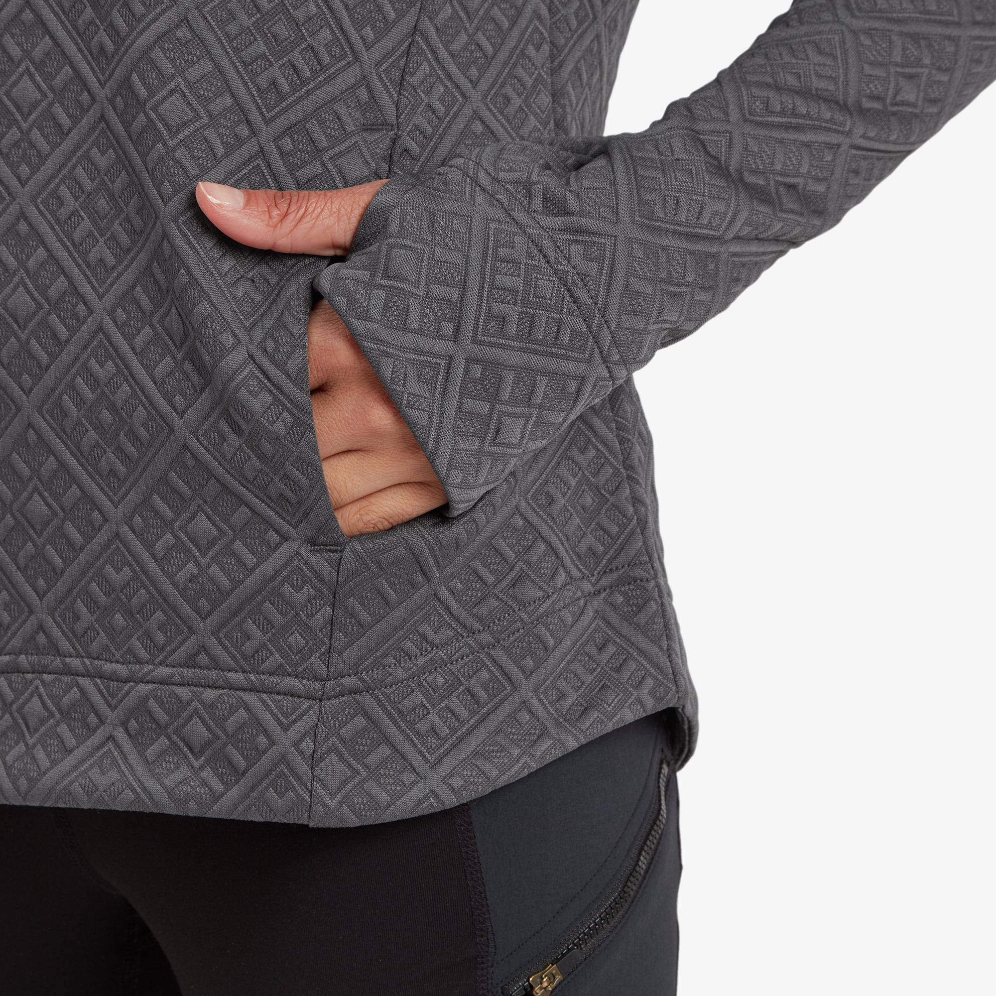 A close-up shot of the model’s hand in the front pocket of the Sherpa Adventure Gear Nyano Pullover in Grey. The pocket is seamlessly integrated into the design, and the diamond pattern is visible across the sleeve and torso. The sleeve extends slightly over her wrist, with a subtle thumbhole feature.