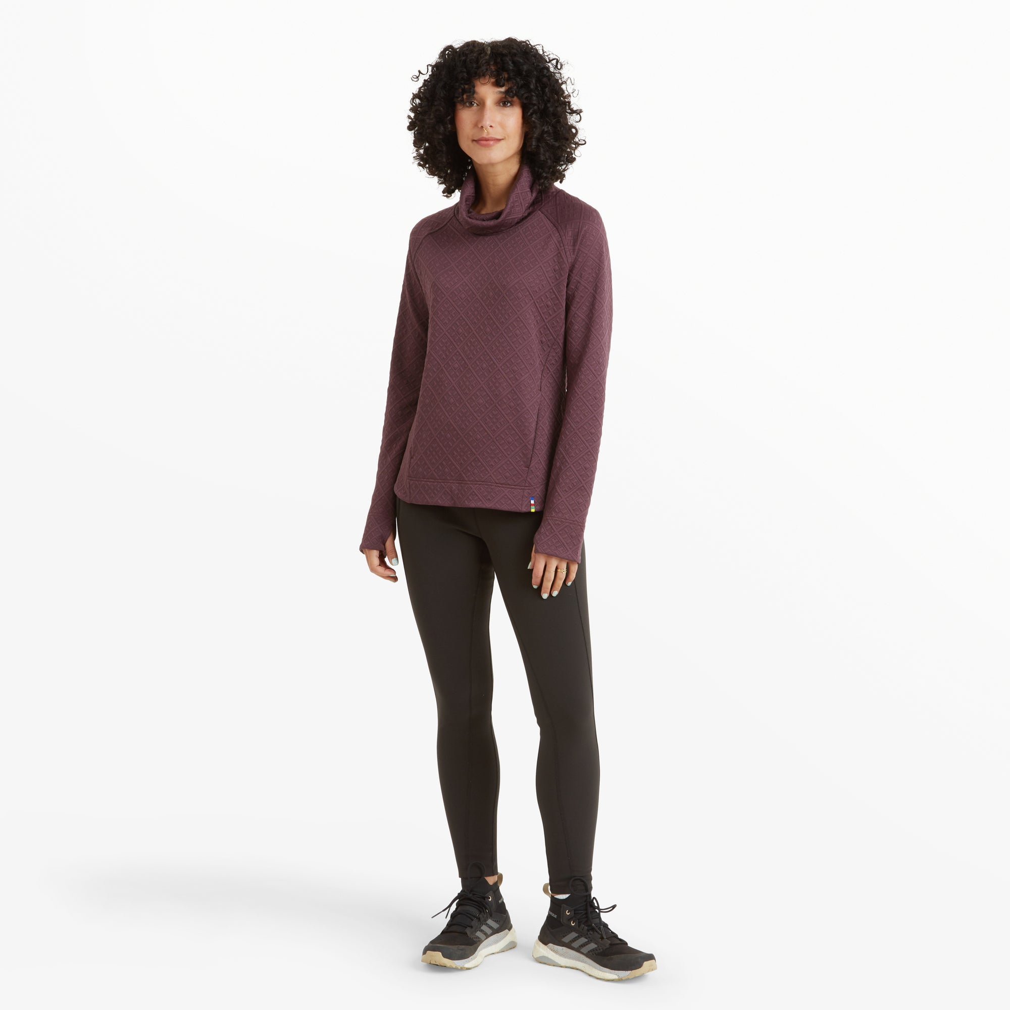 The same woman is shown in a full-body shot wearing the Sherpa Adventure Gear Nyano Pullover in Purple paired with black leggings and black athletic shoes. She stands with a relaxed posture, hands at her sides, against a white background.