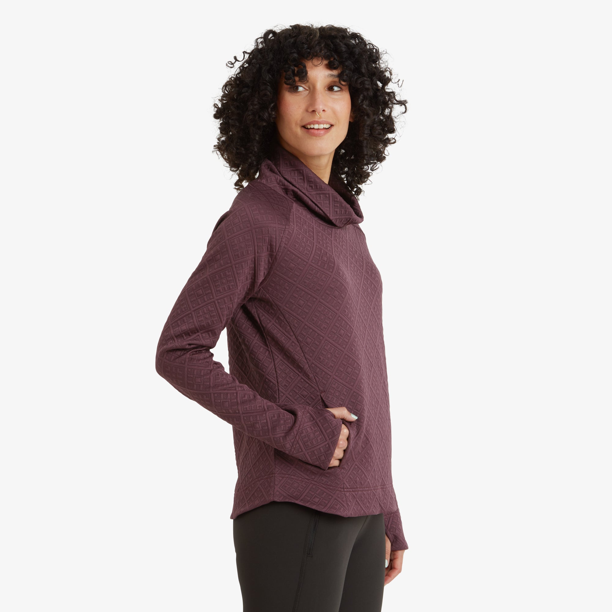 A side profile of the model highlights the draped cowl neck of the Sherpa Adventure Gear Nyano Pullover in Purple and the thumbhole cuffs at the end of the sleeves. She is looking slightly behind her with a subtle smile, her curly hair framing her face.