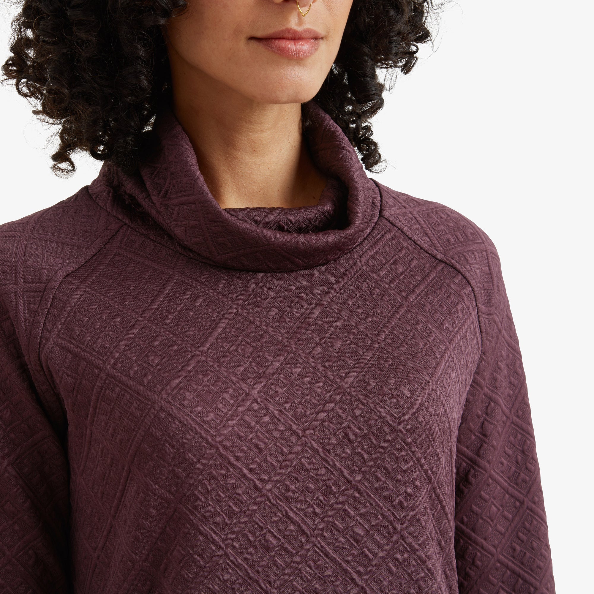 A close-up image of the cowl neck of the Sherpa Adventure Gear Nyano Pullover in Purple, showing the rich texture and the intricate pattern on the fabric. The model’s lips and part of her curly hair are visible.