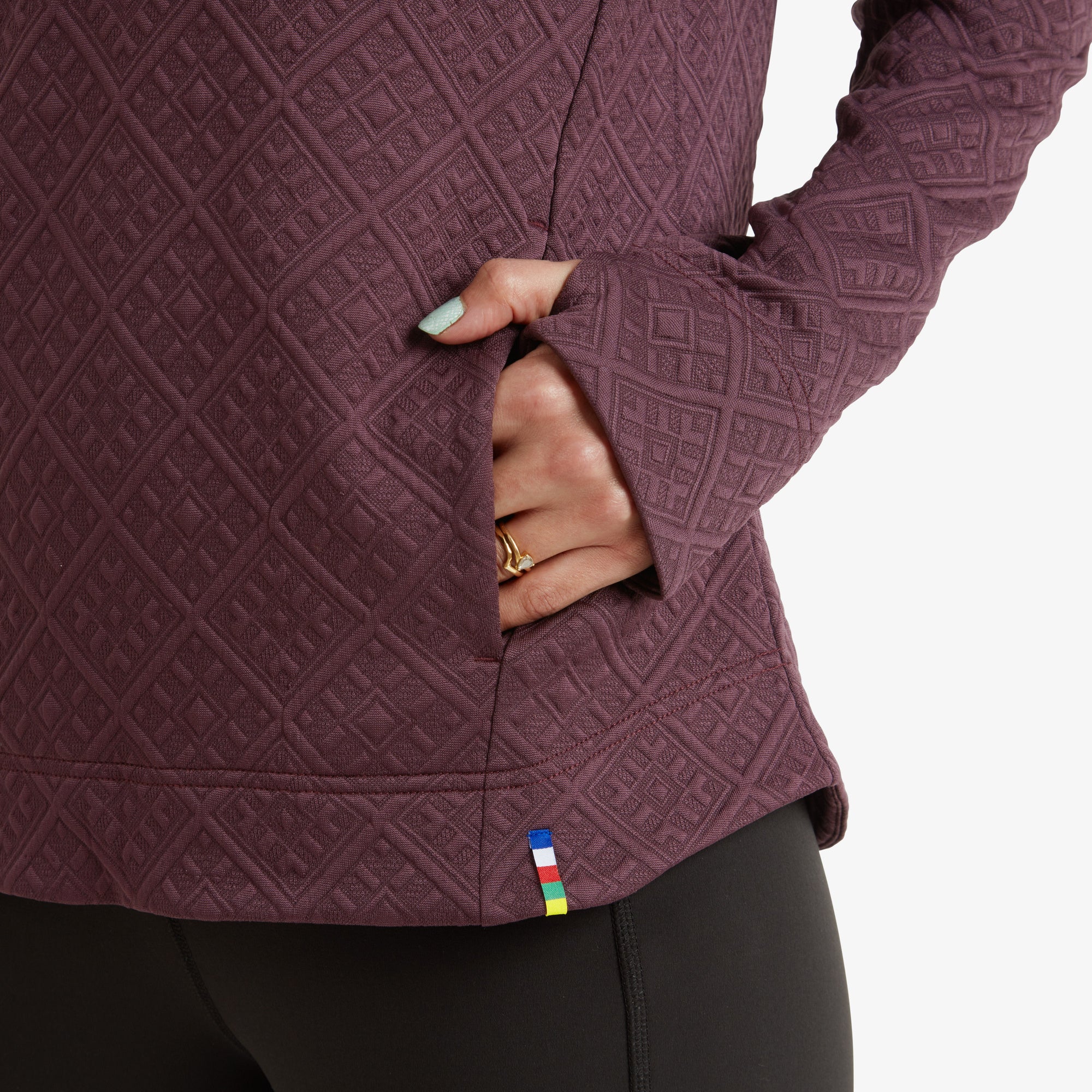 A close-up of the model’s hand resting inside the side pocket of the Sherpa Adventure Gear Nyano Pullover in Purple, highlighting the fabric texture, the small brand tag with multicolored stripes, and the thumbhole feature in the sleeve.