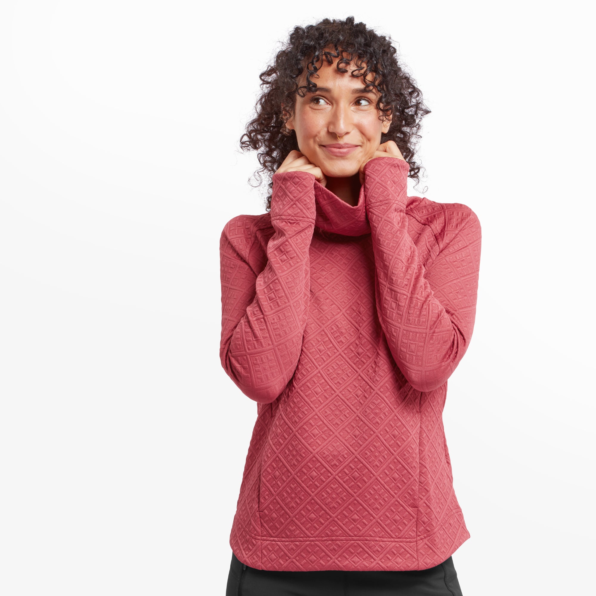 A smiling woman wearing a Sherpa Adventure Gear Nyano Pullover in Red with a relaxed turtleneck design. The pullover features an intricate diamond-shaped quilted pattern, long sleeves, and a comfortable fit. She has her hands playfully tucked under the collar, showcasing the pullover's soft and cozy look.