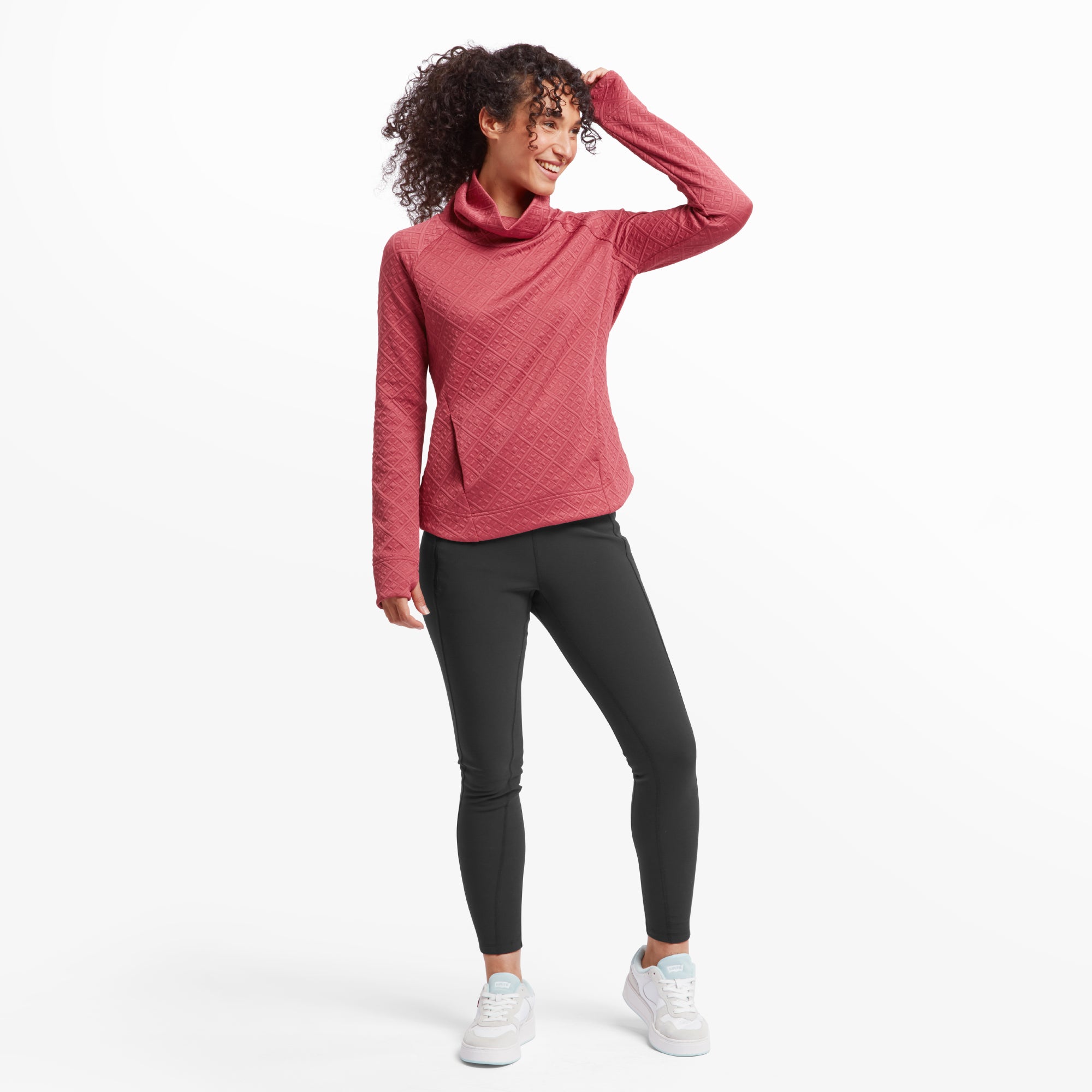 The woman stands confidently, wearing the Sherpa Adventure Gear Nyano Pullover in Red paired with black leggings and white trainers. She has one hand on her head, radiating a casual, stylish vibe. The pullover's slightly curved hemline and flattering fit are visible, emphasising its versatile design for everyday wear.