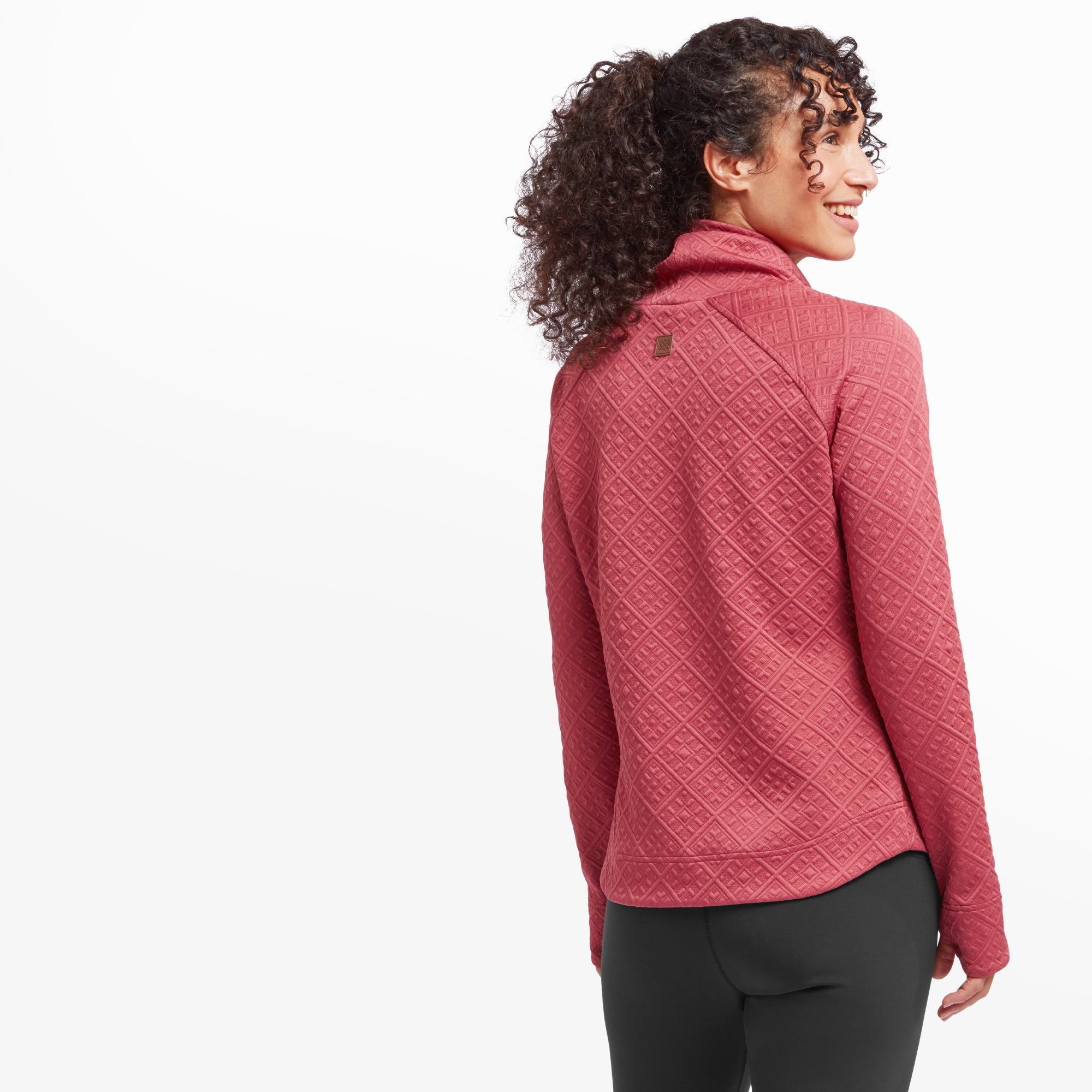 A rear view of the woman showcasing the back of the Sherpa Adventure Gear Nyano Pullover in Red. The textured diamond pattern extends across the entire garment, and a small logo detail is present at the back of the neck. Her hair is tied in a high ponytail, drawing attention to the relaxed turtleneck.