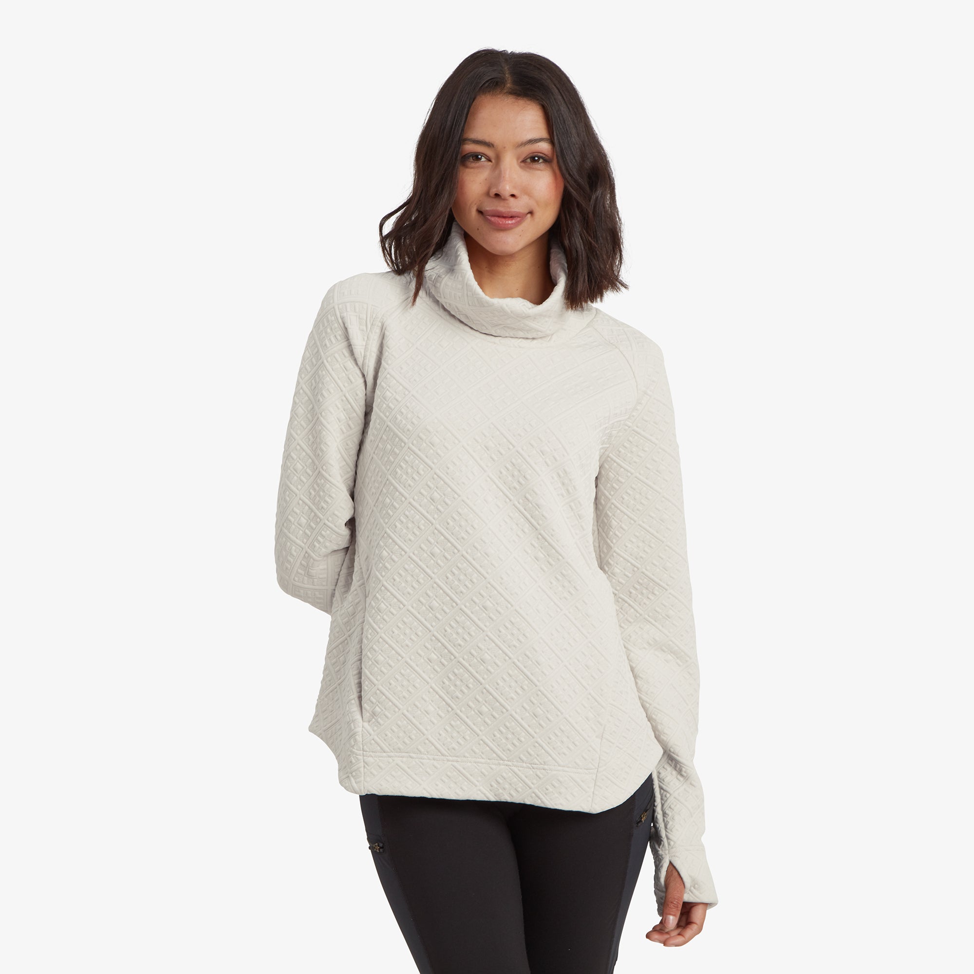 A woman wears a Sherpa Adventure Gear Nyano Pullover in White with a cozy cowl neck, slightly smiling. The quilted pattern has diamond-shaped detailing, creating a textured and soft appearance. She pairs it with black leggings, posing front-facing with relaxed hands.