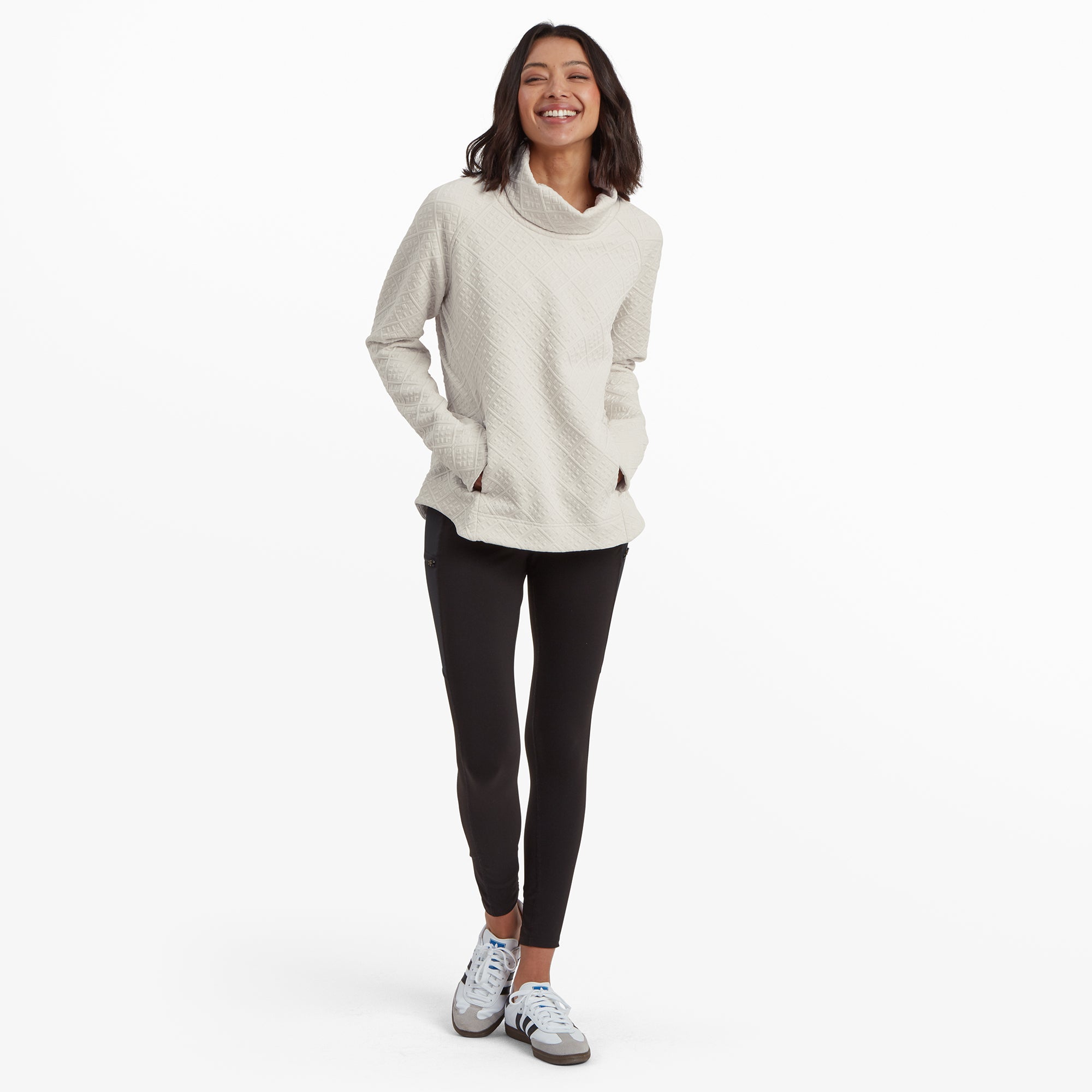 The same woman showcases a full-body view of the Sherpa Adventure Gear Nyano Pullover in White. She stands confidently, hands in the pullover's side pockets, paired with fitted black leggings and sporty trainers. Her wide smile and relaxed posture highlight the pullover's casual, comfortable fit.