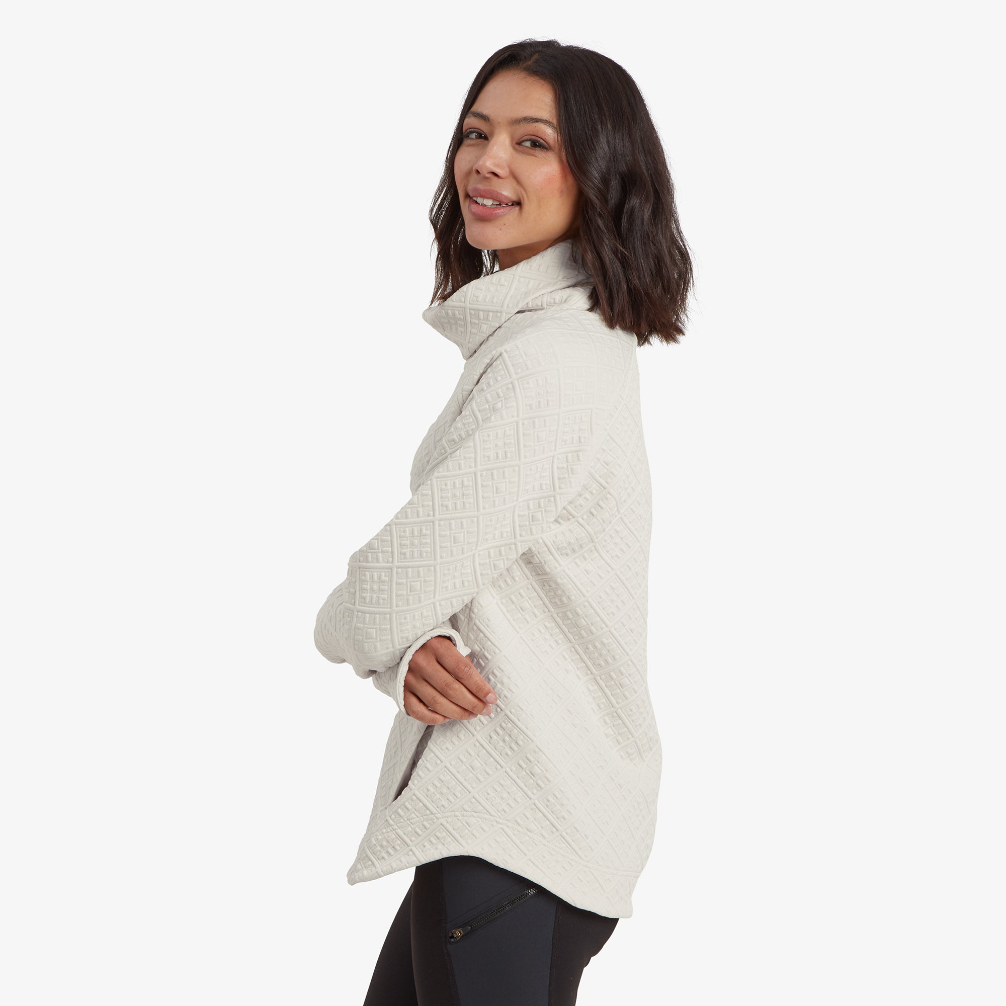 A side profile of the woman in the Sherpa Adventure Gear Nyano Pullover in White, showcasing its relaxed fit and high-low hemline. The raised cowl neck is emphasised, and her hands are tucked casually into the pullover's side pockets. The quilting pattern is prominently visible along the side.