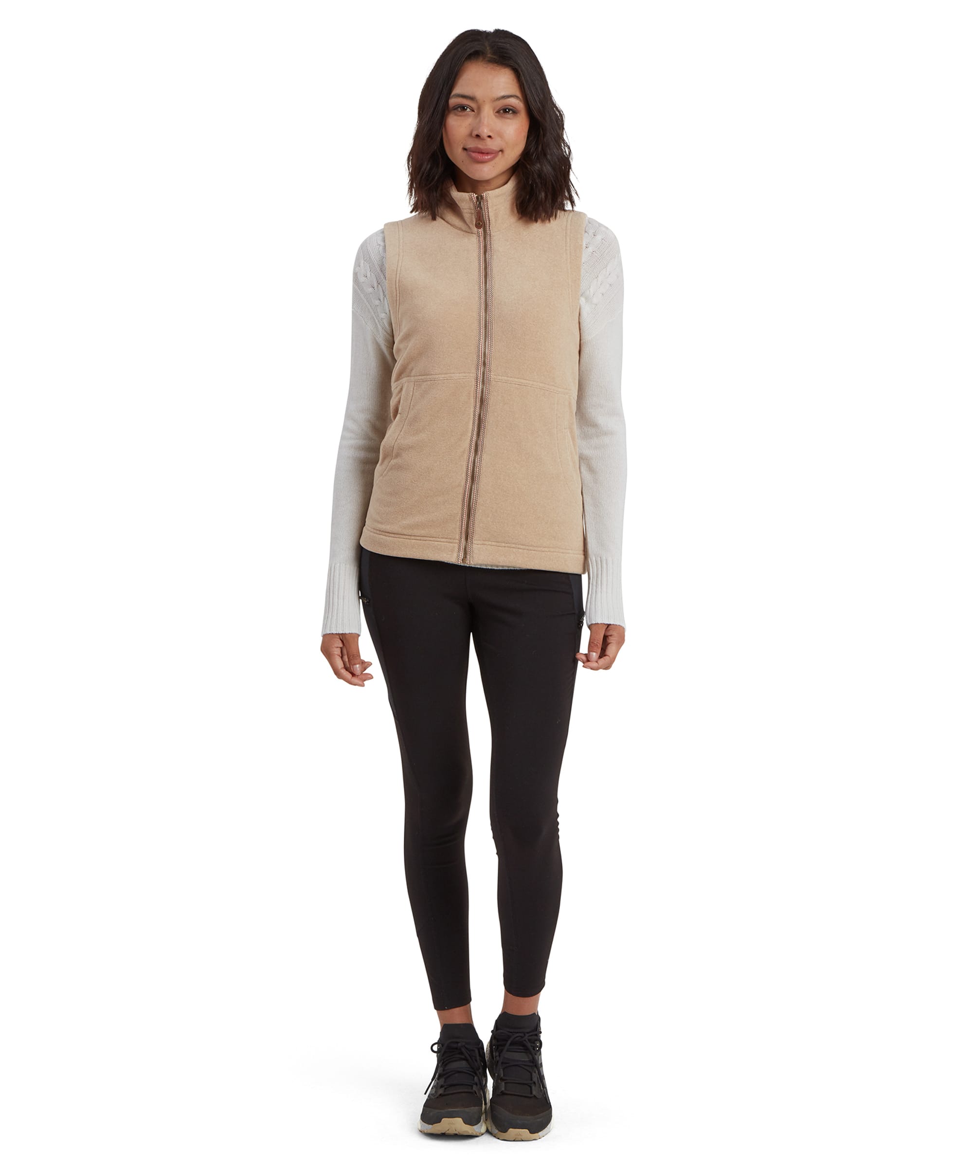 The same woman models the full outfit, standing in a relaxed pose. The Sherpa Adventure Gear Rolpa Eco Vest in Beige is paired with black leggings and athletic shoes, illustrating its versatility for casual or outdoor settings. The neutral tones complement the overall ensemble.