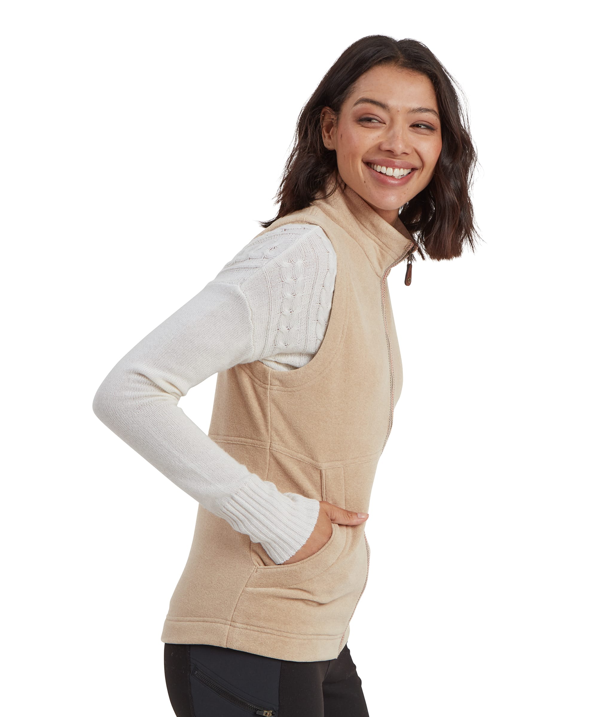 A side profile of the model as she smiles, highlighting the snug fit and tailored design of the Sherpa Adventure Gear Rolpa Eco Vest in Beige. The fleece material appears soft and cozy, with functional hand pockets for added utility.