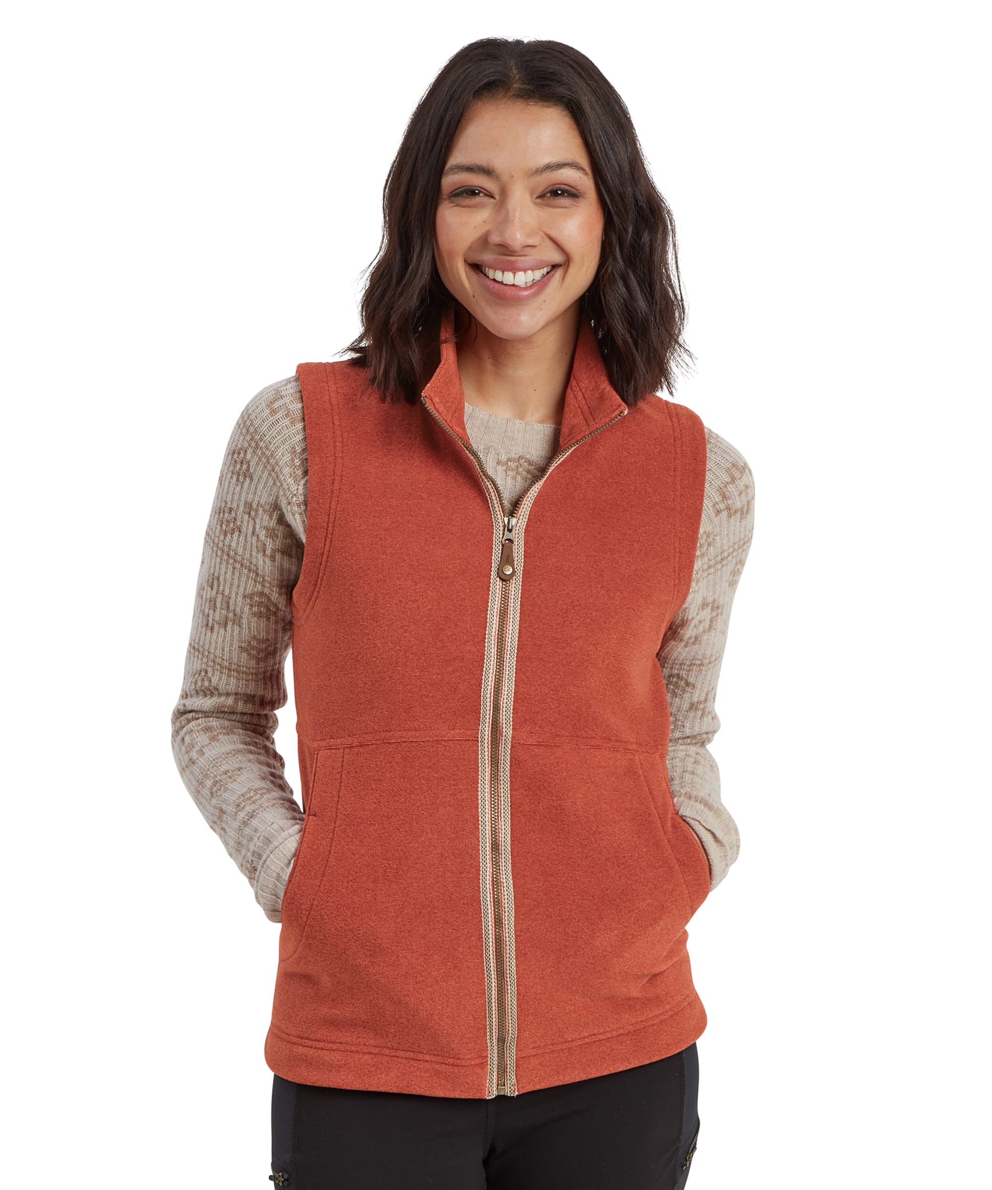 A woman smiling while wearing a Sherpa Adventure Gear Rolpa Eco Vest in Brown. The gilet has a front zipper and side pockets, and she pairs it with a beige, patterned long-sleeve shirt and black leggings. The image showcases the front design of the gilet.