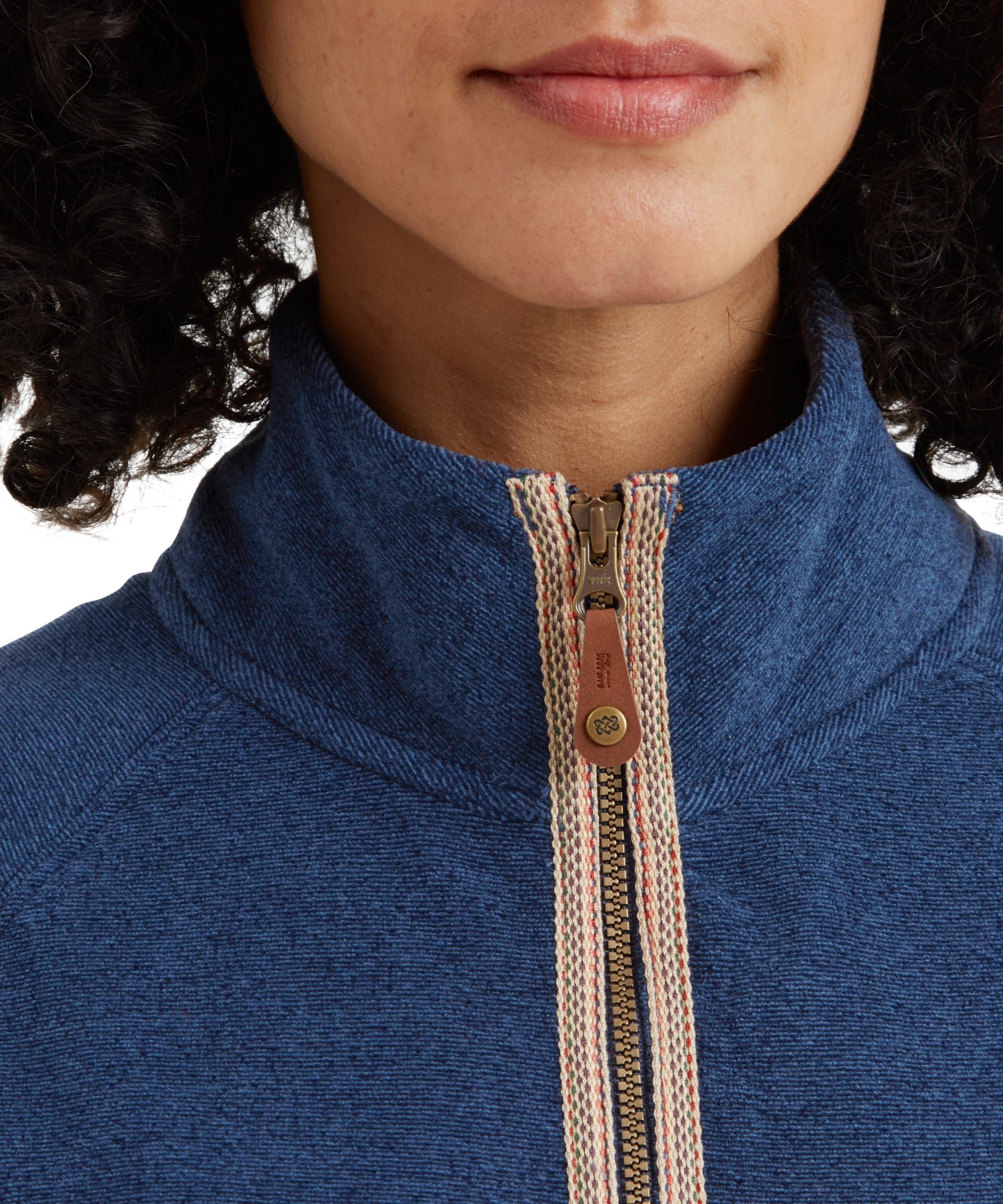 A close-up of the high collar and full-length zipper of the Sherpa Adventure Gear Rolpa Eco Jacket in Blue. The zipper features a tan and white woven trim with a small leather pull tab that has a circular embossed logo.