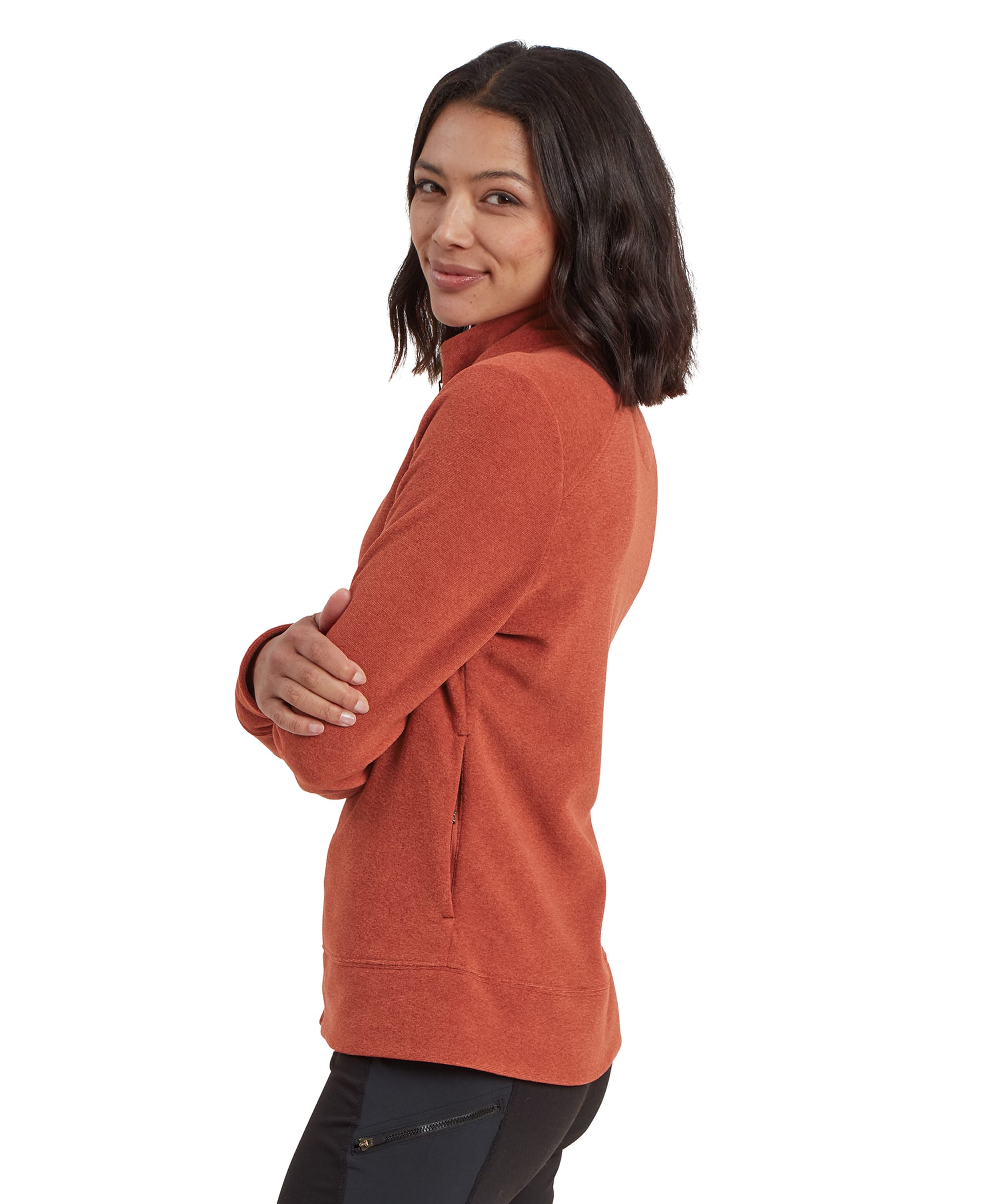 A side view of the woman wearing the Sherpa Adventure Gear Rolpa Eco Jacket in Brown, smiling while turning slightly toward the camera. Her arms are crossed in front of her chest, giving a better view of the side seam and one of the jacket's zippered pockets. The soft fleece texture of the jacket is visible, and the relaxed fit allows for layering underneath.