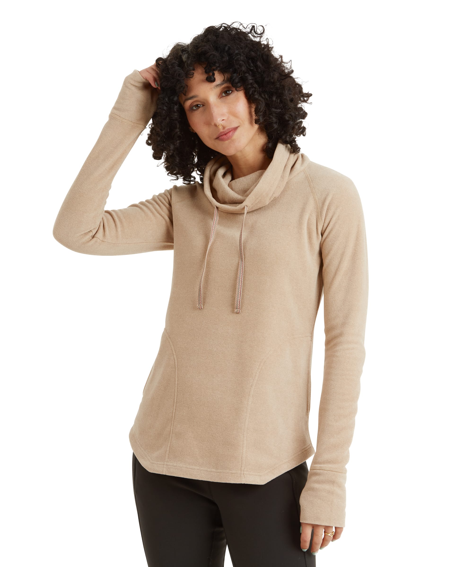 A woman wearing a Sherpa Adventure Gear Rolpa Eco Pullover in Beige with a relaxed fit. The top features a cowl neck with drawstrings, thumb holes, and a subtle curved hemline. She has her hand lightly resting on her head, showcasing the soft and cozy material of the pullover, paired with black leggings.