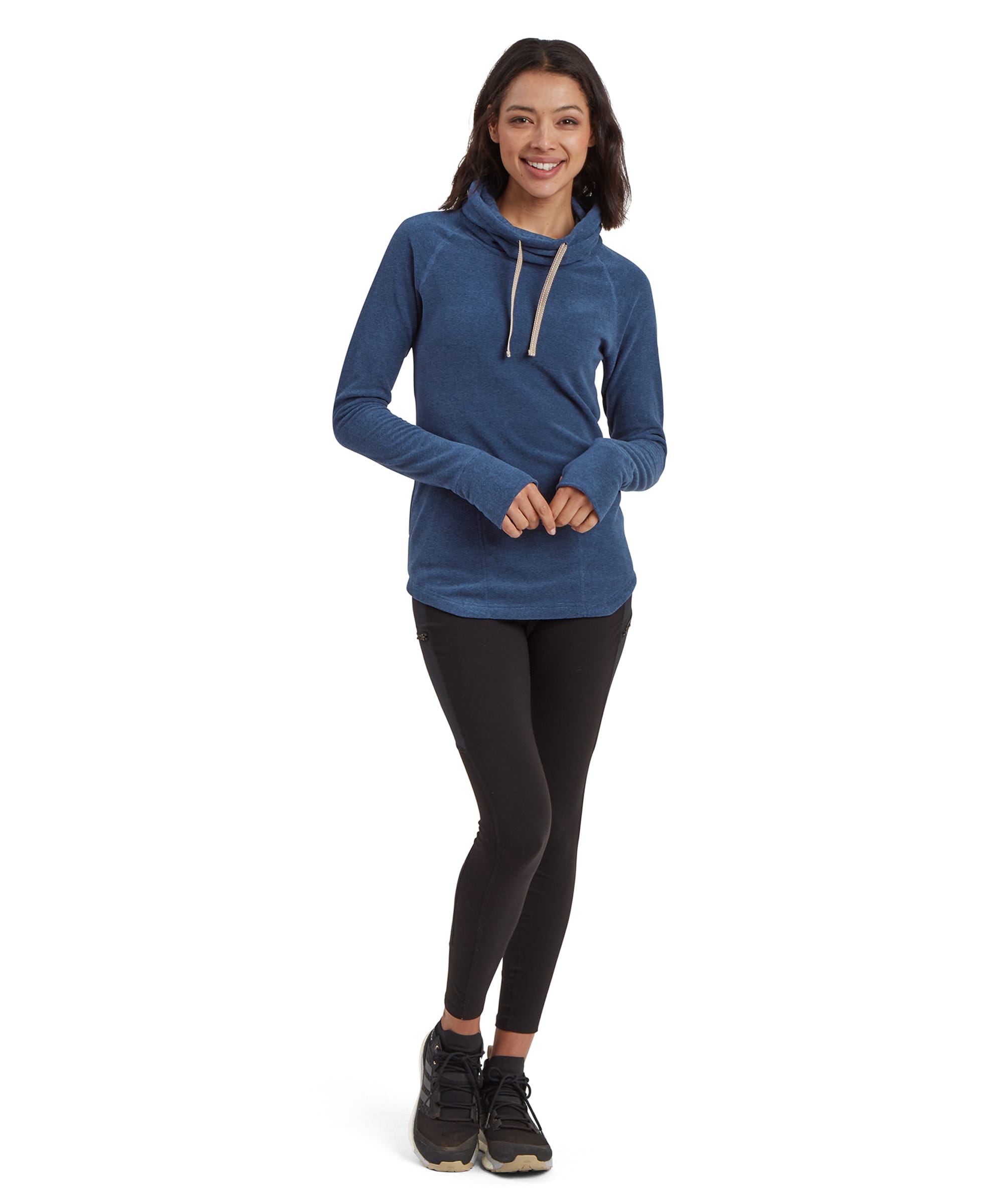 A full-body shot of the same woman wearing the Sherpa Adventure Gear Rolpa Eco Pullover in Blue paired with black leggings and black trainers. She stands casually with one foot slightly forward and hands relaxed in front of her. The high cowl neck and thumb holes on the sleeves are clearly visible. The background is plain white, emphasising the outfit.