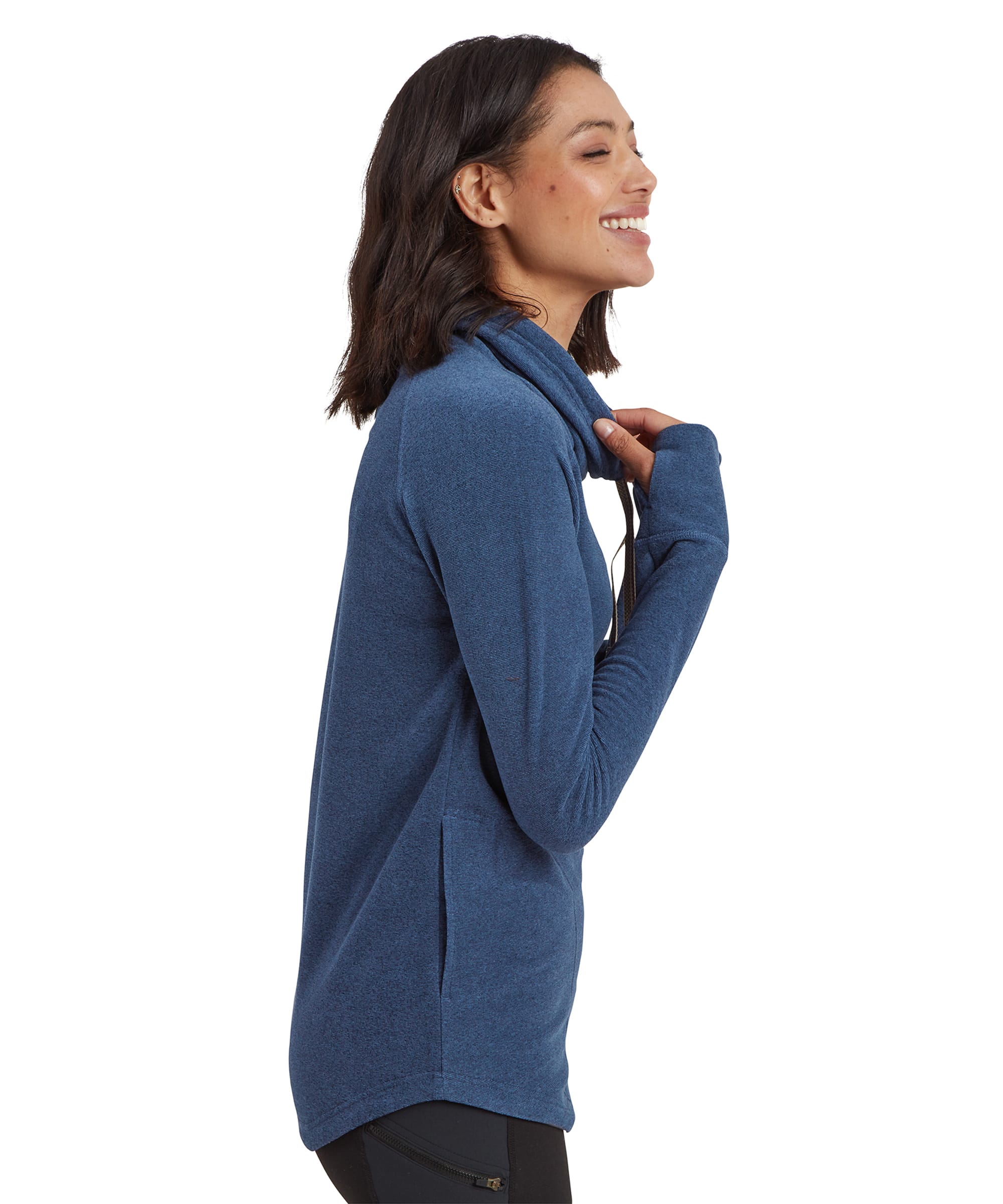 A side profile view of the woman wearing the Sherpa Adventure Gear Rolpa Eco Pullover in Blue. She is smiling and slightly pulling at the collar, showcasing the high neckline and drawstrings. The fabric appears soft, and the sweater has a slightly curved hemline. Her body faces the side, while her head is turned slightly toward the camera.