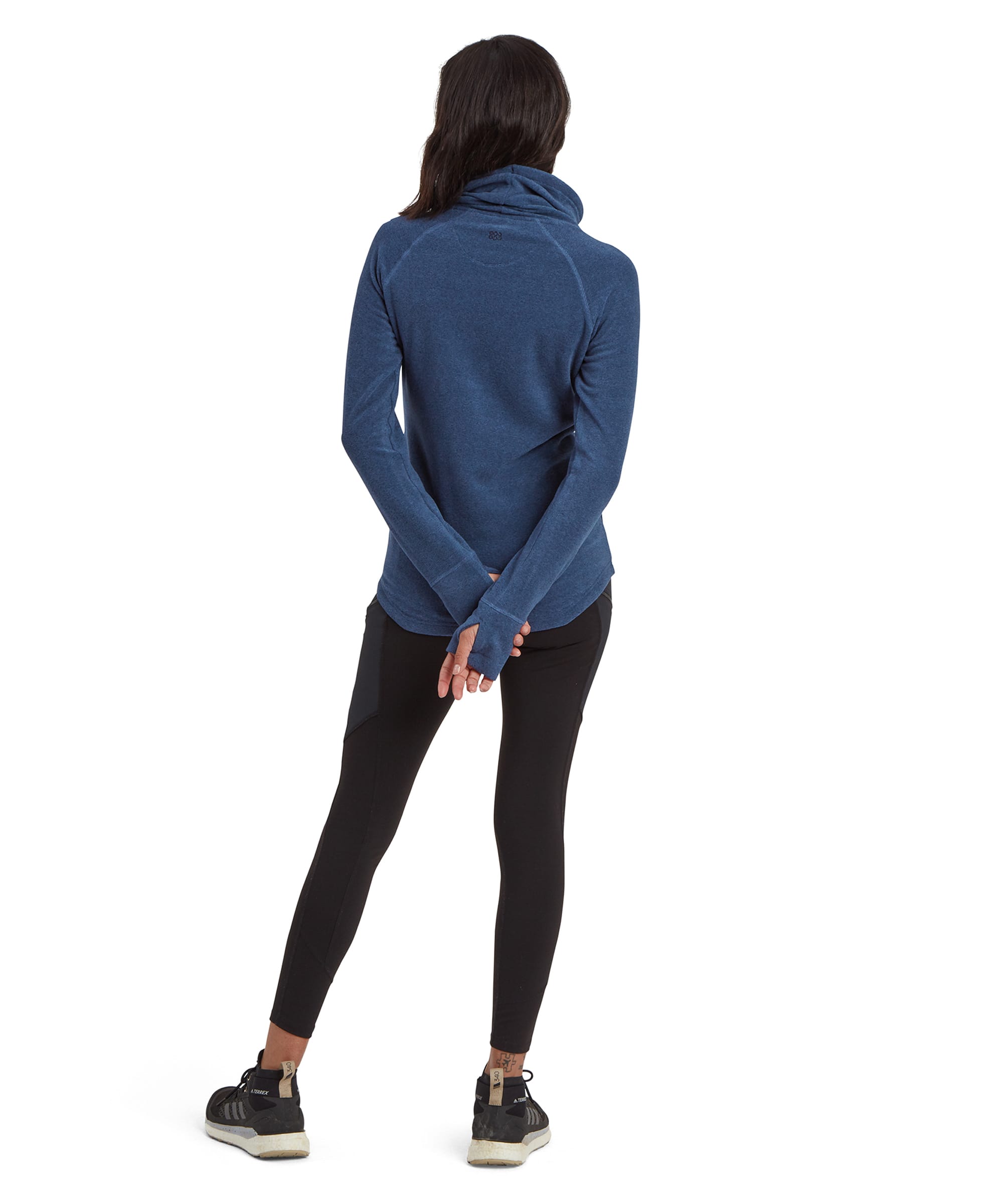 A rear view of the woman wearing the Sherpa Adventure Gear Rolpa Eco Pullover in Blue. The back of the pullover is plain blue, with a seam running across the upper back area and extending to the sides. The sweater slightly covers her hips, and the sleeves extend down to her hands, showing the thumb holes. She is wearing black leggings and black shoes.