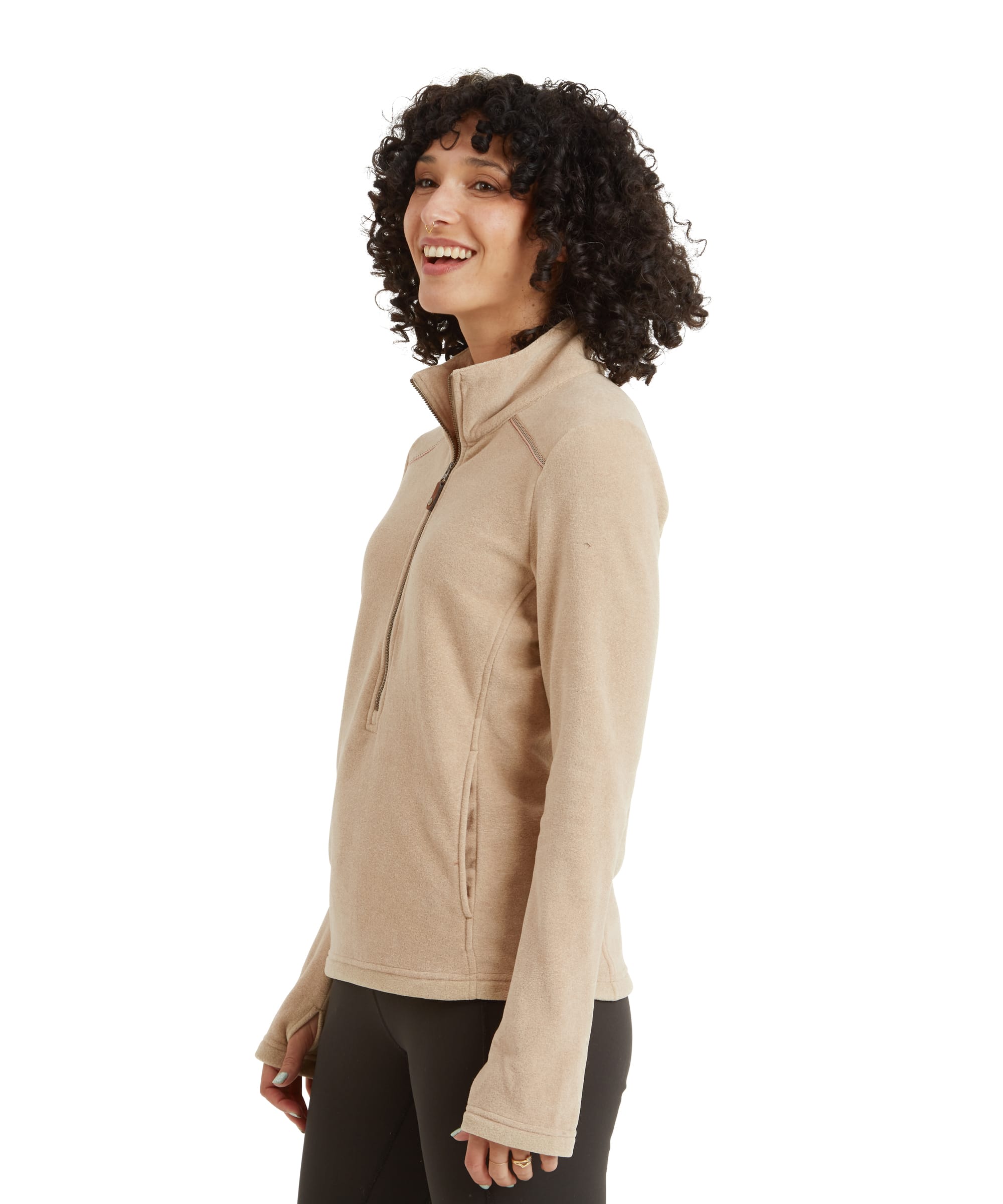 A side view of the woman wearing the Sherpa Adventure Gear Rolpa Eco Pullover Zip in Beige. The zipper is slightly unzipped, highlighting the high neckline, and the side seams provide a tailored fit. She is smiling, with her hands loosely by her sides.