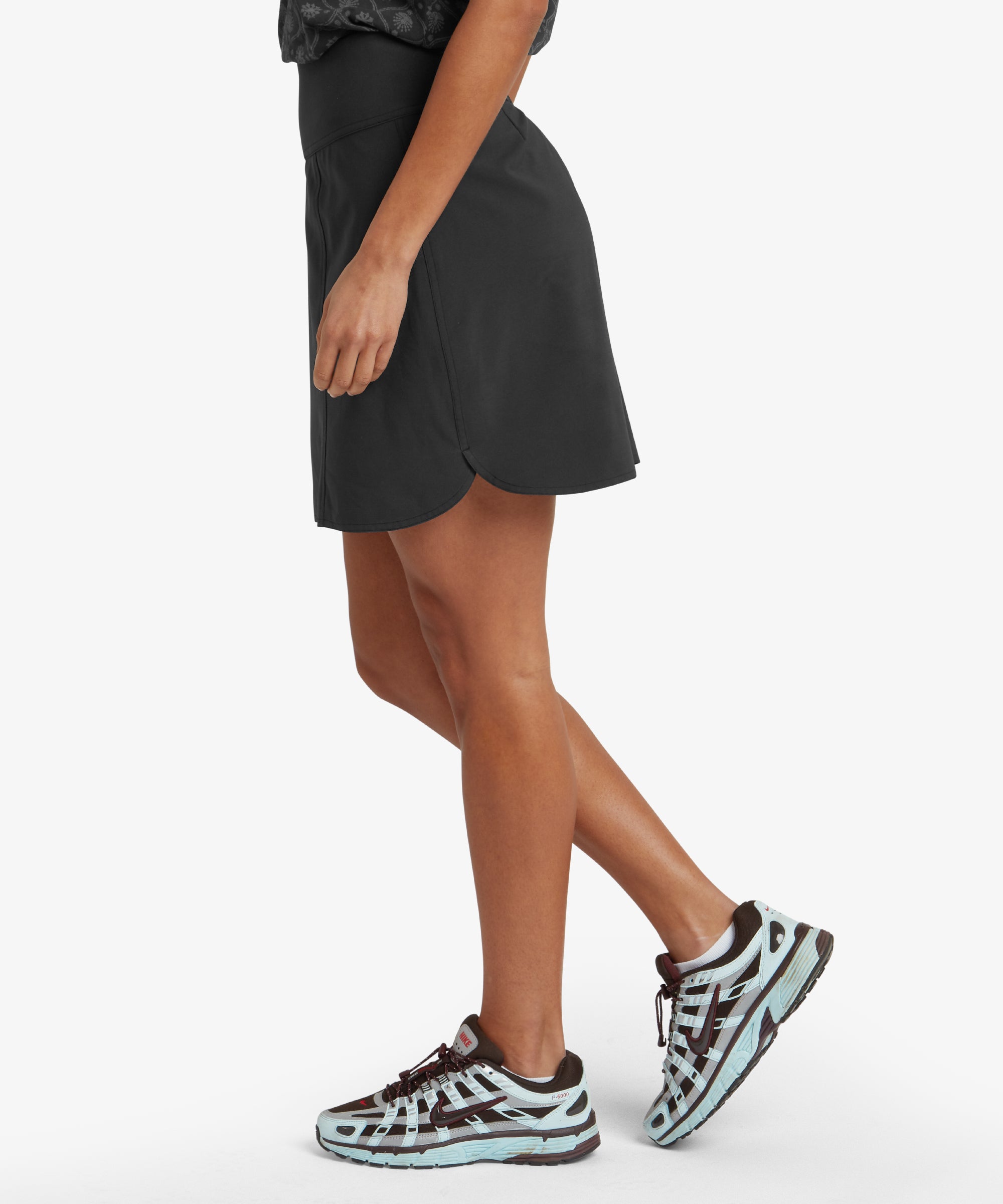 A side view of a woman wearing a Sherpa Adventure Gear Sajilo Adventure Skort in Black. The skort has a smooth and structured design with a slight slit on the side for comfort. She is paired with a light green t-shirt and white trainers, standing on a neutral background.