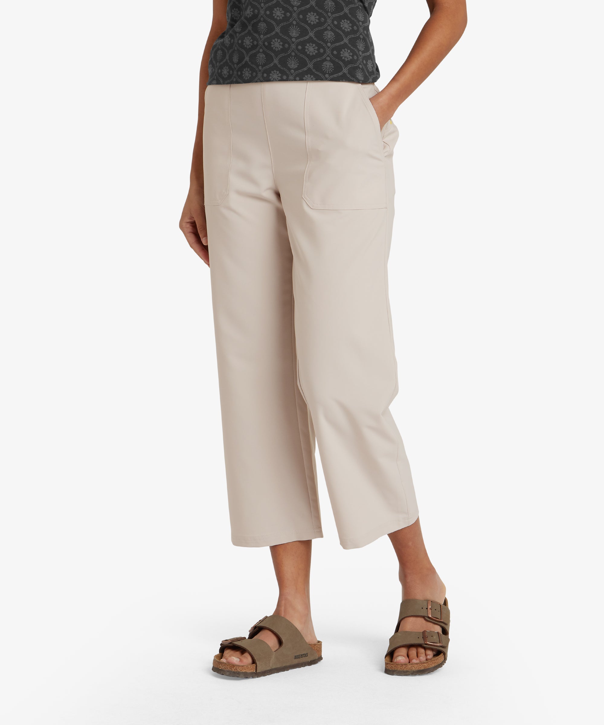 A close-up view of a model's lower body wearing Sherpa Adventure Gear Sajilo Capri in Beige paired with white trainers. The pants have a straight-leg cut and a cropped hem that falls above the ankle. The model's stance is relaxed with one foot slightly forward.