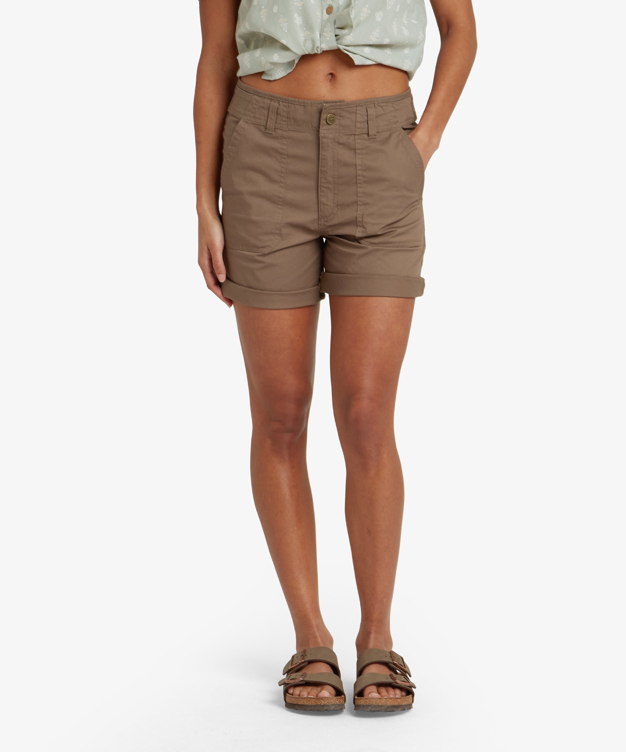 A woman modeling the Sherpa Adventure Gear Tenjing Shorts in Grey. The shorts have a relaxed fit, with a rolled cuff and a front button closure with a zipper fly. The shorts feature two front pockets, subtle stitching detail, and belt loops at the waistband. She is wearing a light green, printed button-up shirt tied at the waist and brown sandals.