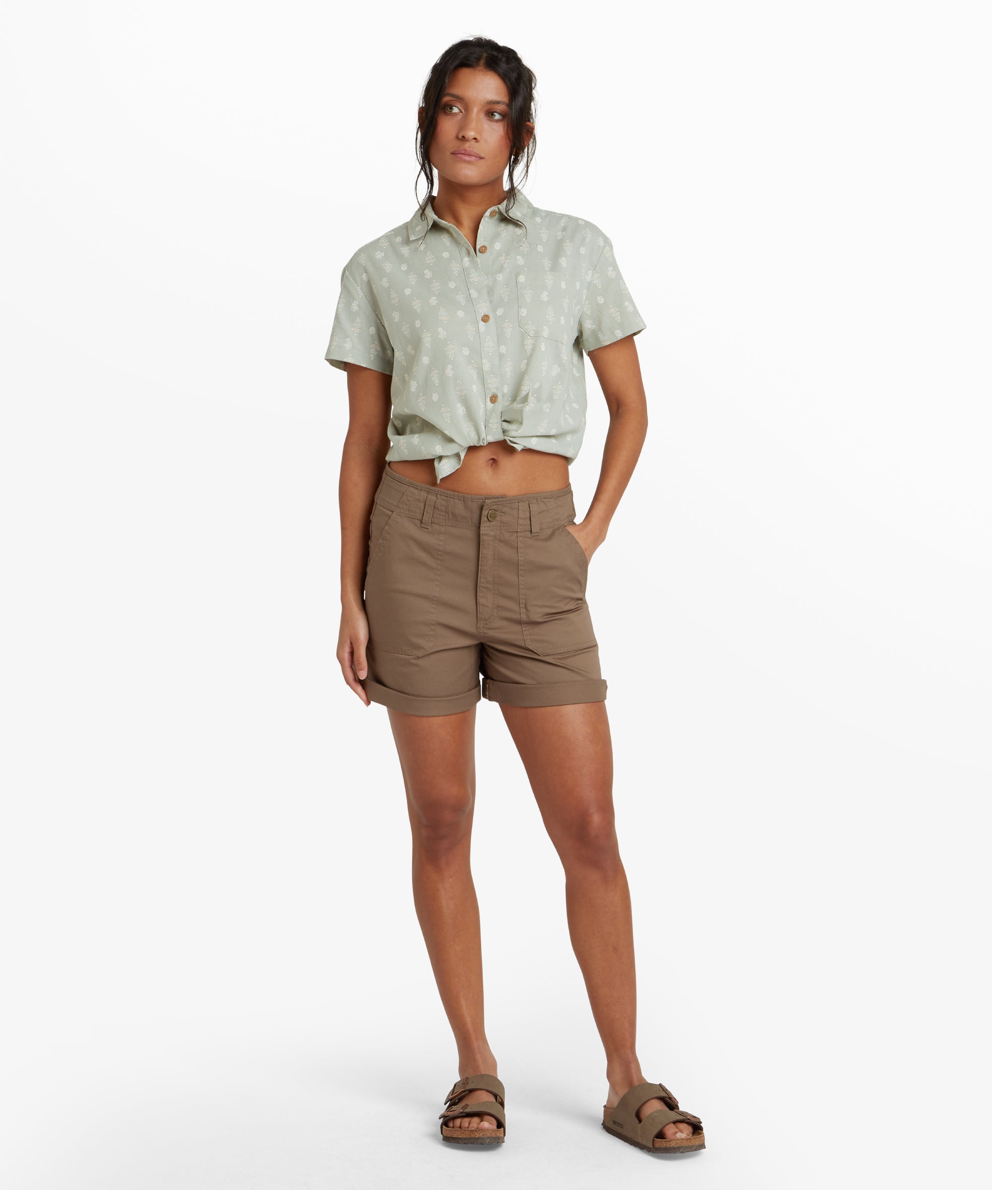 Full-body view of a woman wearing the Sherpa Adventure Gear Tenjing Shorts in Grey. She is styled with a short-sleeve, collared, green print shirt tied at the front and casual brown sandals. She stands with one hand in her pocket, highlighting the shorts' practical and versatile design.