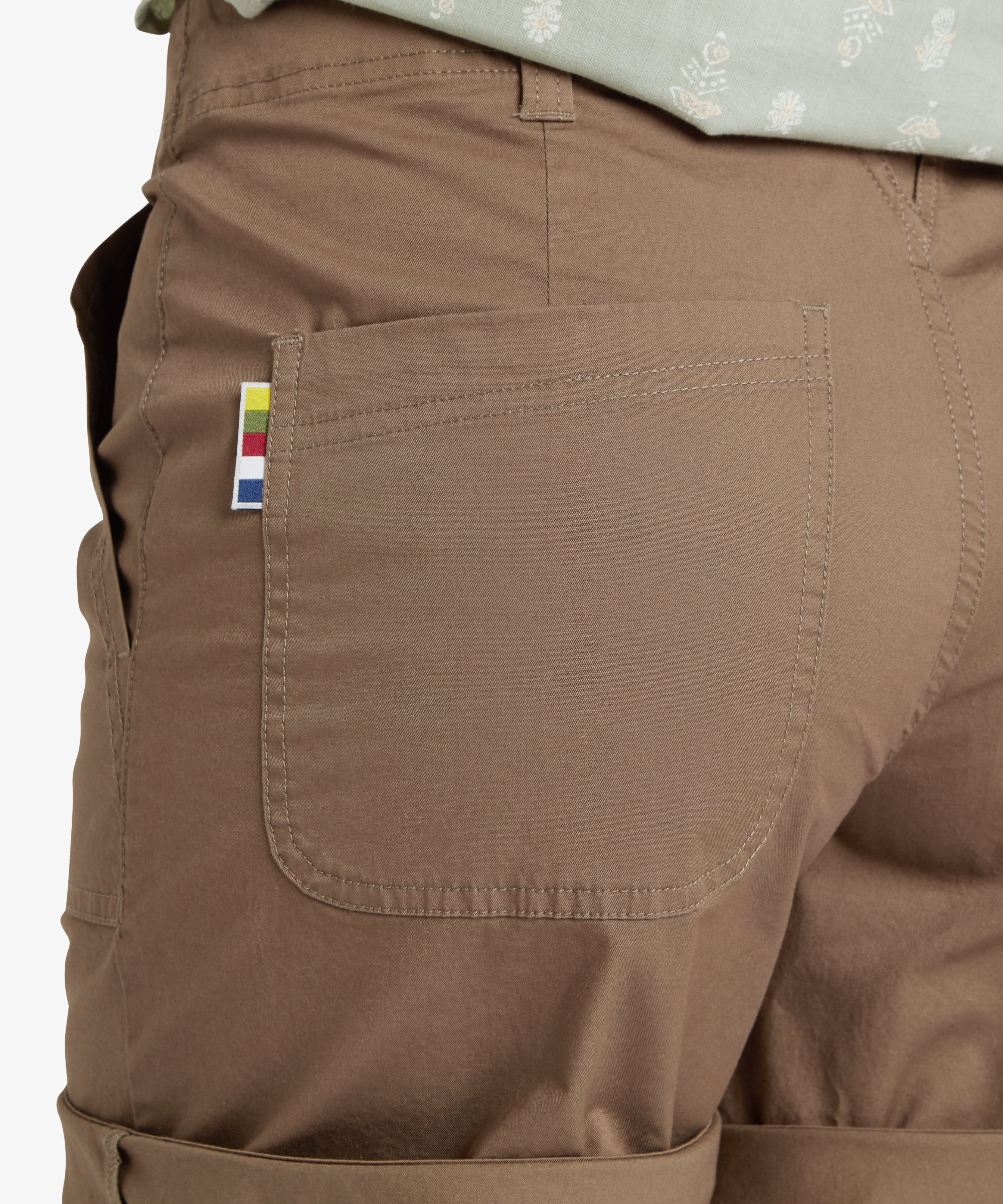 Detailed close-up of the back pocket on the Sherpa Adventure Gear Tenjing Shorts in Grey. The patch pocket features reinforced stitching, and a small rectangular multicolored woven logo tag is attached to the outer side of the pocket, adding a subtle branded touch.