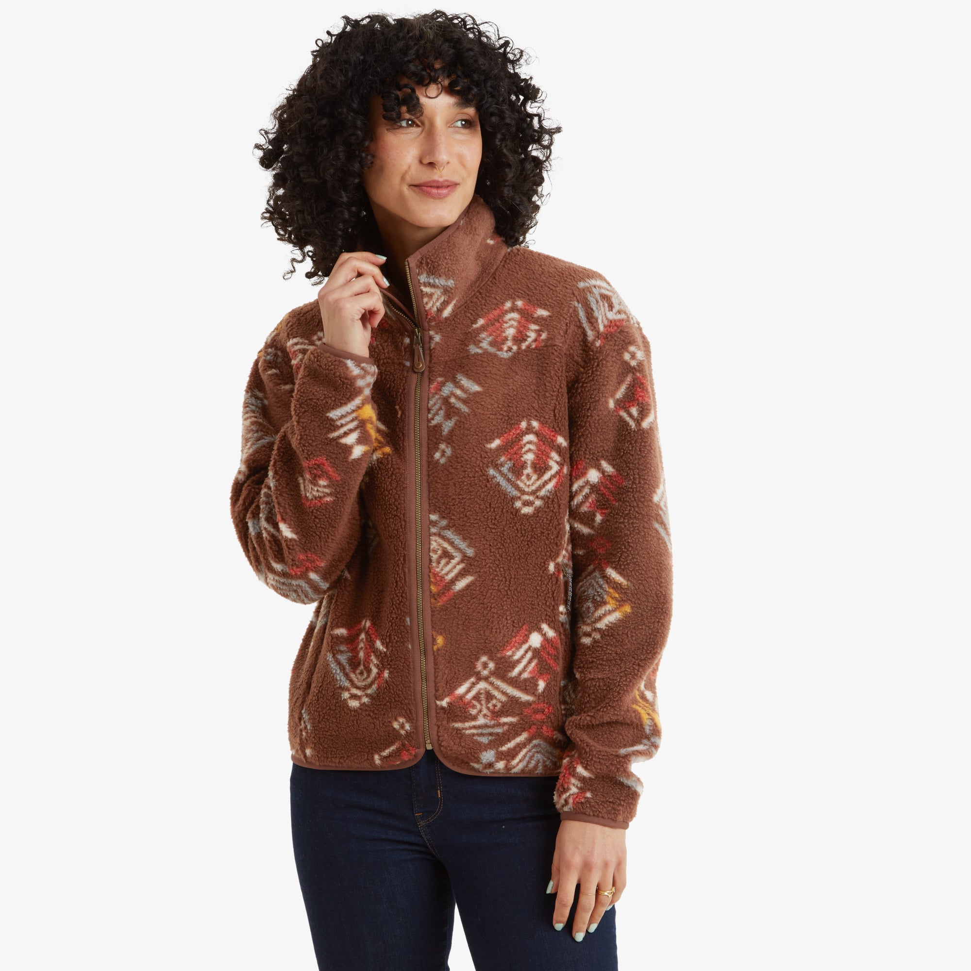 A woman wearing a Sherpa Adventure Gear Uddesya Eco Jacket in Brown featuring colorful geometric patterns. The jacket has a high collar and full zip closure, with the model gently holding the collar and smiling slightly. She is styled in dark blue skinny jeans.