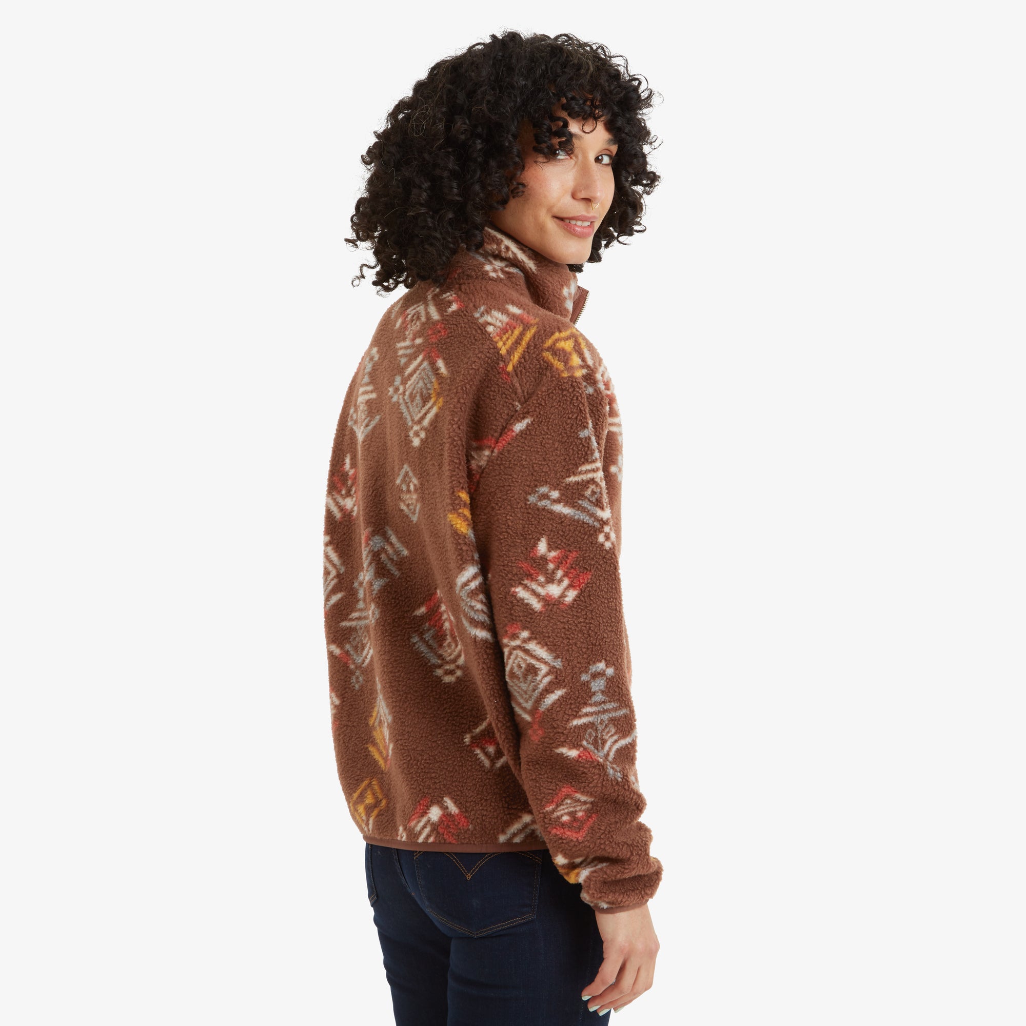 The model showcases the rear side of the Sherpa Adventure Gear Uddesya Eco Jacket in Brown, looking back over her shoulder with a subtle smile. The intricate geometric patterns continue seamlessly across the back.