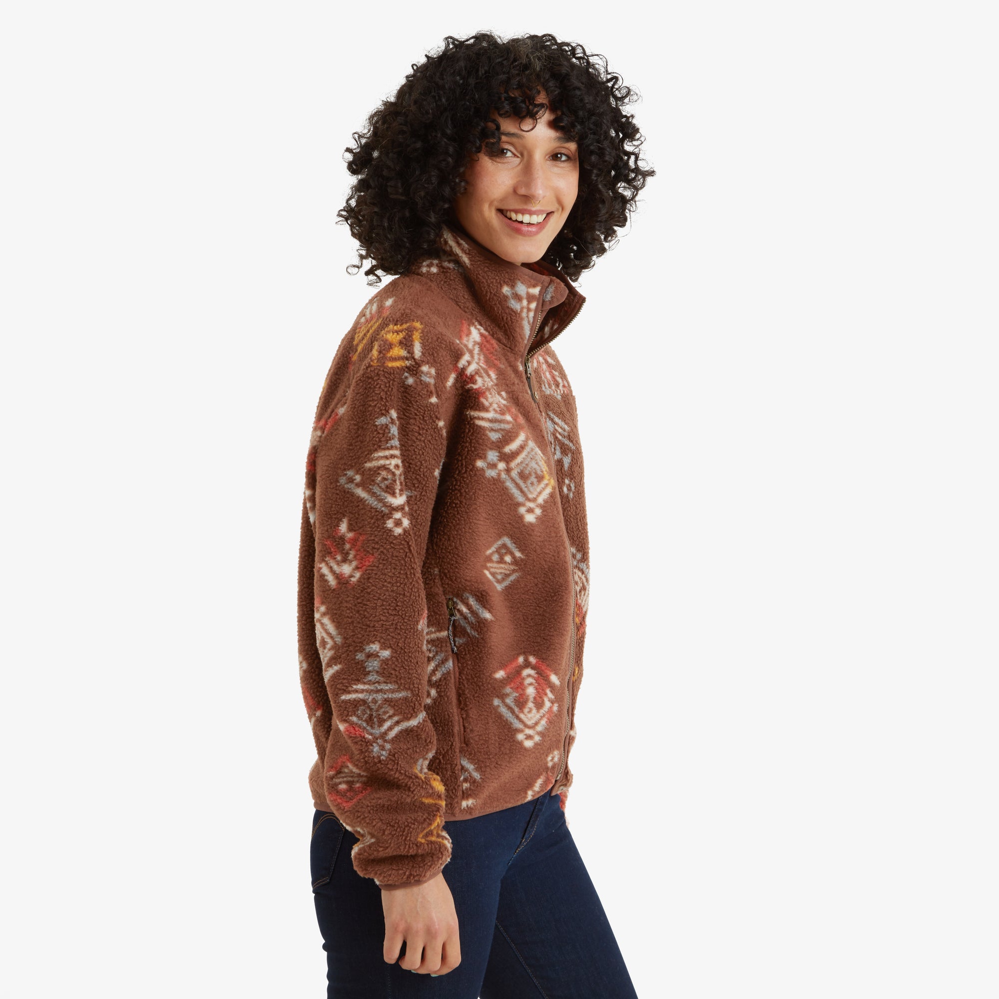 The woman smiles warmly, showing the side profile of the Sherpa Adventure Gear Uddesya Eco Jacket in Brown. The high collar is zipped up, and the colorful geometric print is visible along the arms and body.