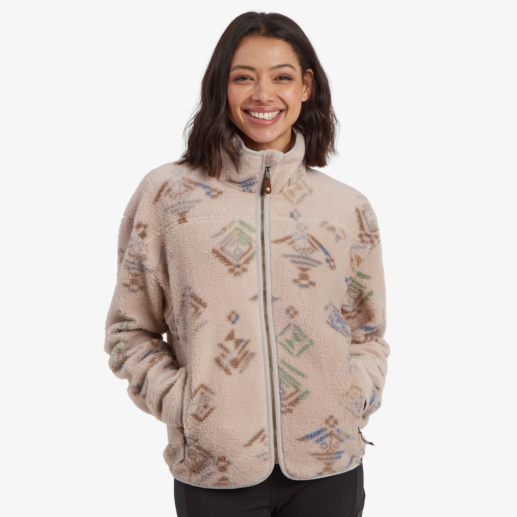 A close-up front-facing image of a woman wearing a Sherpa Adventure Gear Uddesya Eco Jacket in Light Brown with colorful geometric patterns. The jacket has a soft fleece texture, a high collar, and a visible zipper in the center. The model is smiling warmly with her hands in the jacket pockets.