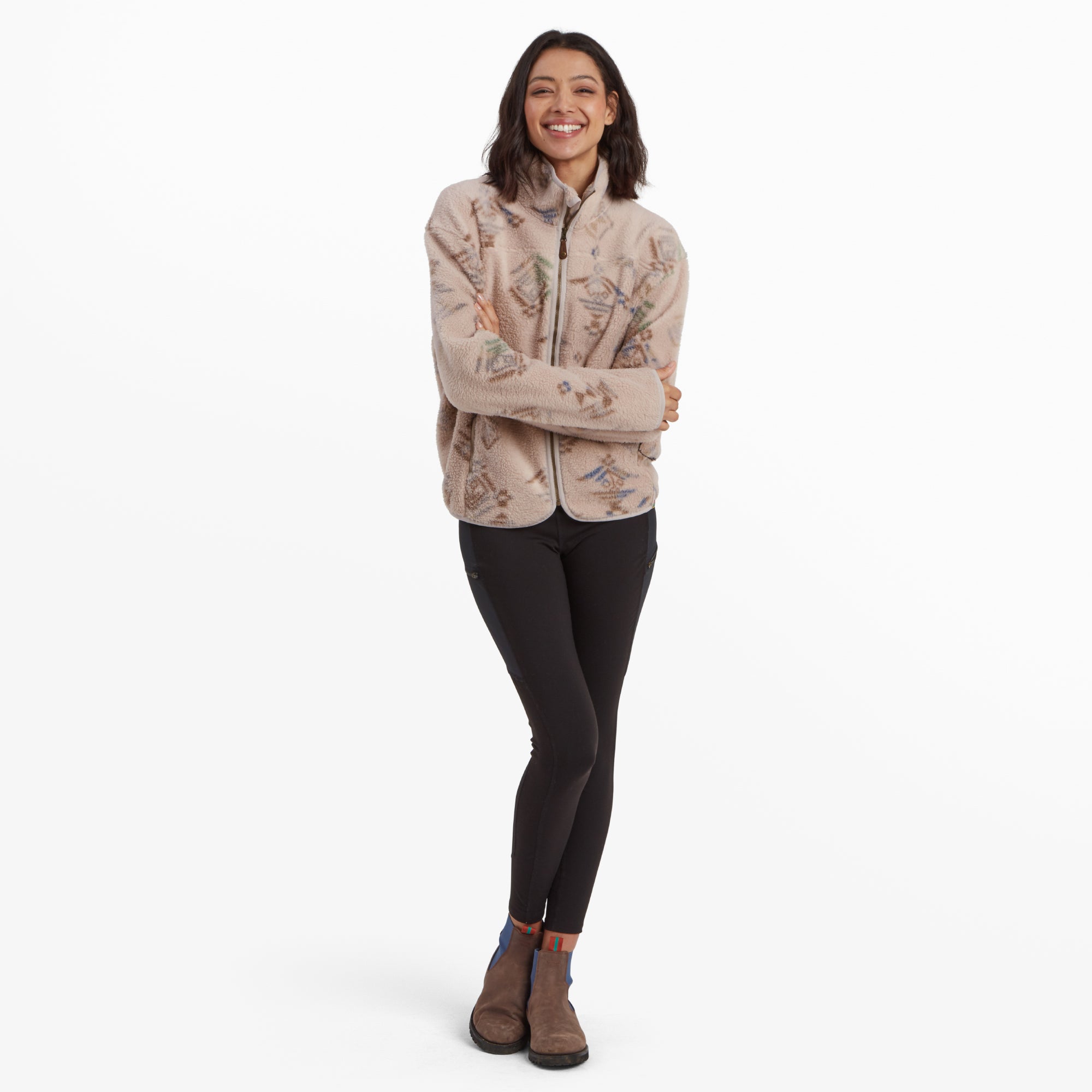 A full-body view of the model wearing the same Sherpa Adventure Gear Uddesya Eco Jacket in Light Brown paired with black leggings and brown ankle boots. The model is smiling and standing with her arms crossed, showcasing a casual and comfortable look.