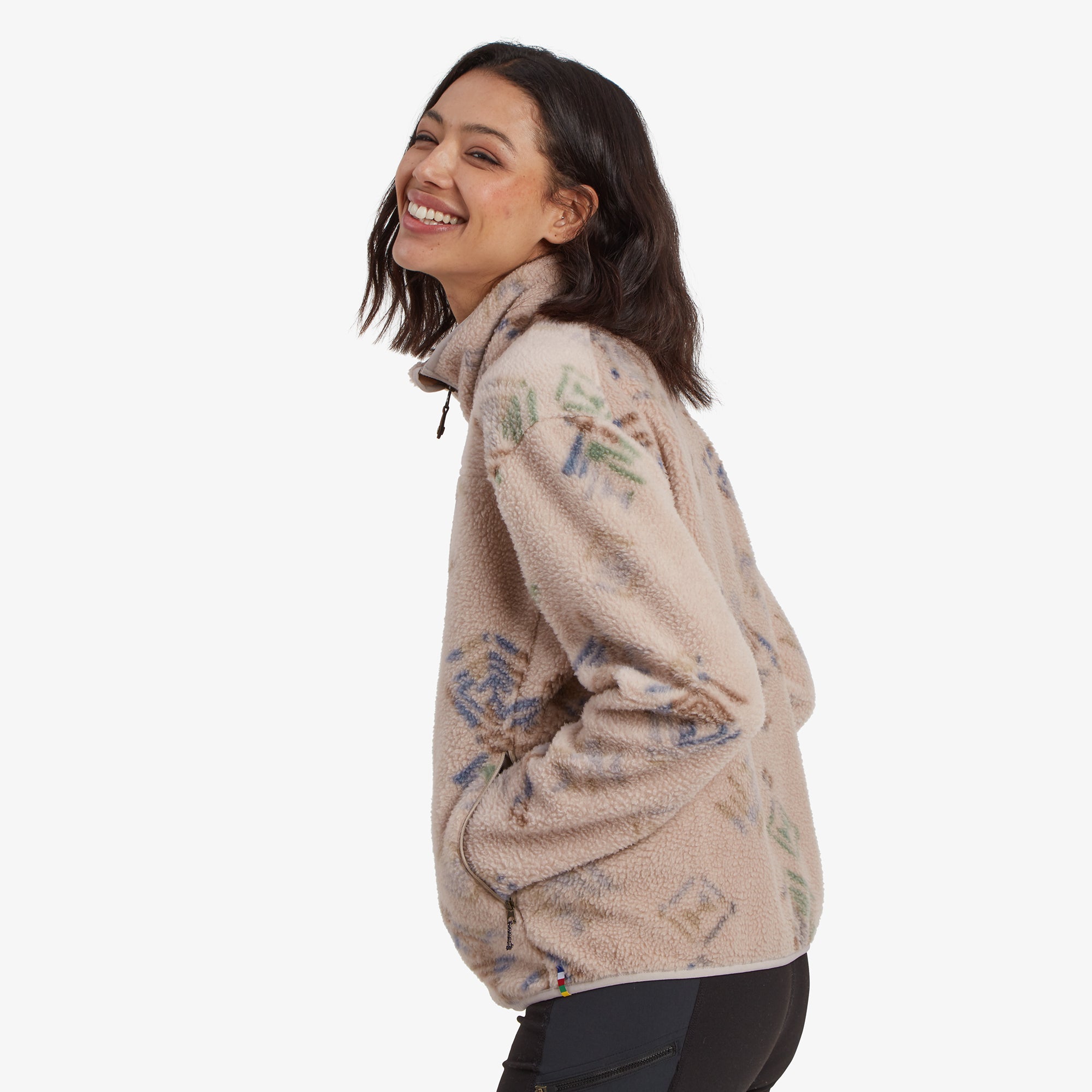 A side profile of the model wearing the Sherpa Adventure Gear Uddesya Eco Jacket in Light Brown, slightly turned and looking over her shoulder with a cheerful smile. The fleece material and colorful geometric patterns are clearly visible on the side of the jacket.