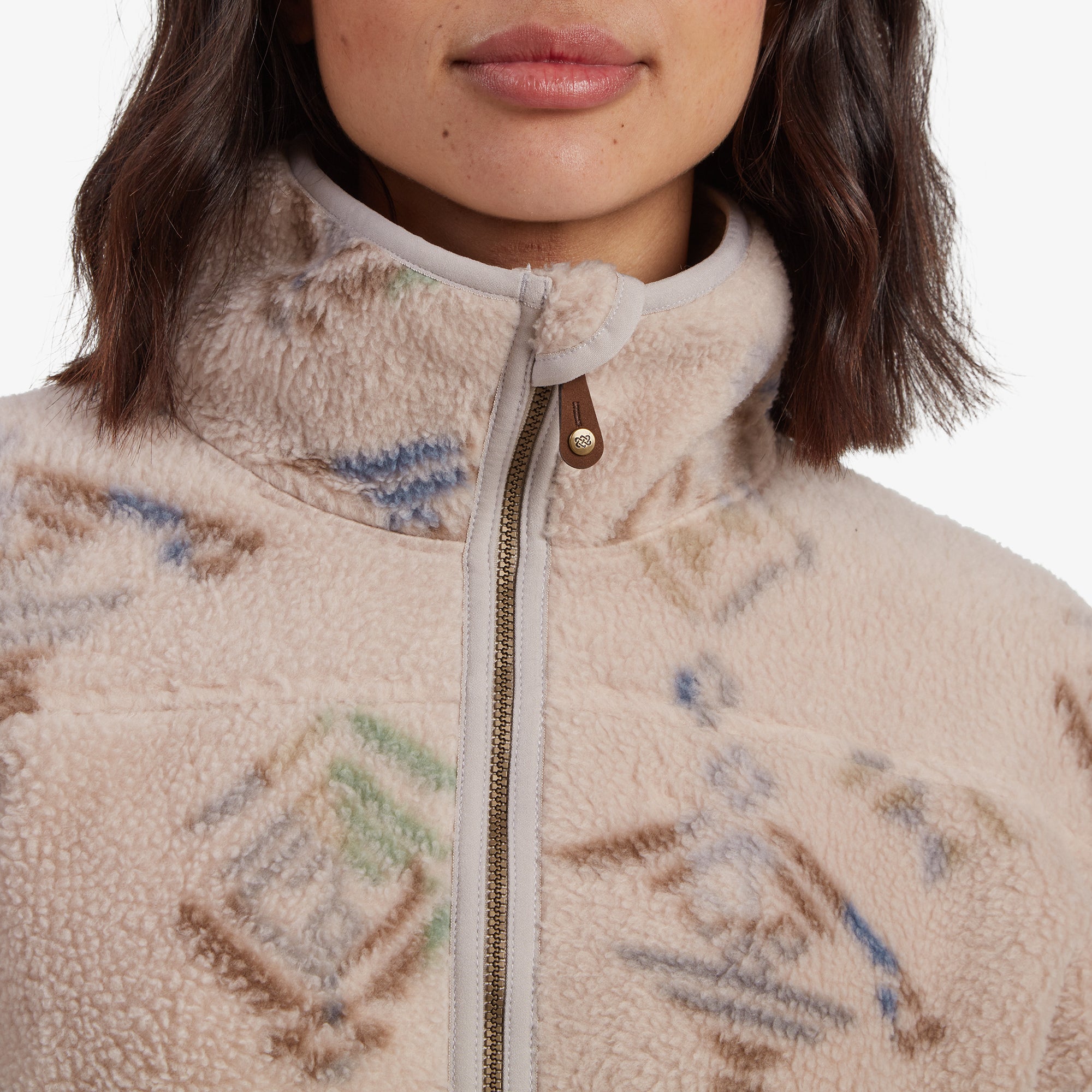 A close-up of the high collar and zippered front of the Sherpa Adventure Gear Uddesya Eco Jacket in Light Brown, highlighting the detail of the stitching, pattern, and zipper pull. The geometric pattern in soft shades of blue, green, and brown stands out on the fleece material.