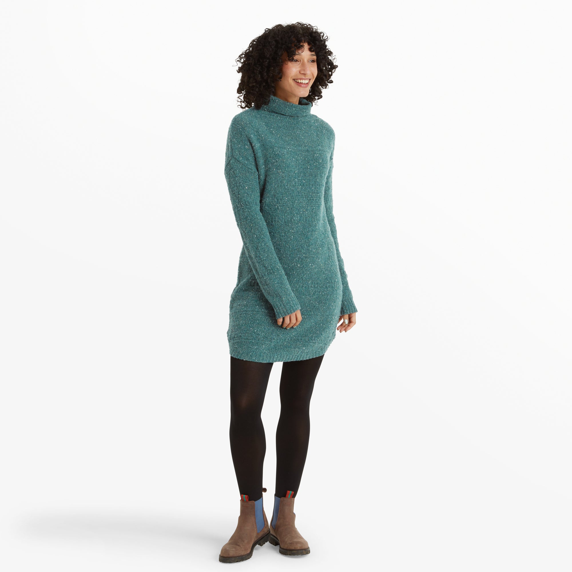 A woman poses in a Sherpa Adventure Gear Yuden Dress in Blue with a turtleneck design, standing front-facing. The dress has a speckled texture, paired with black tights and brown Chelsea boots featuring blue elastic panels. She smiles, holding a relaxed posture.