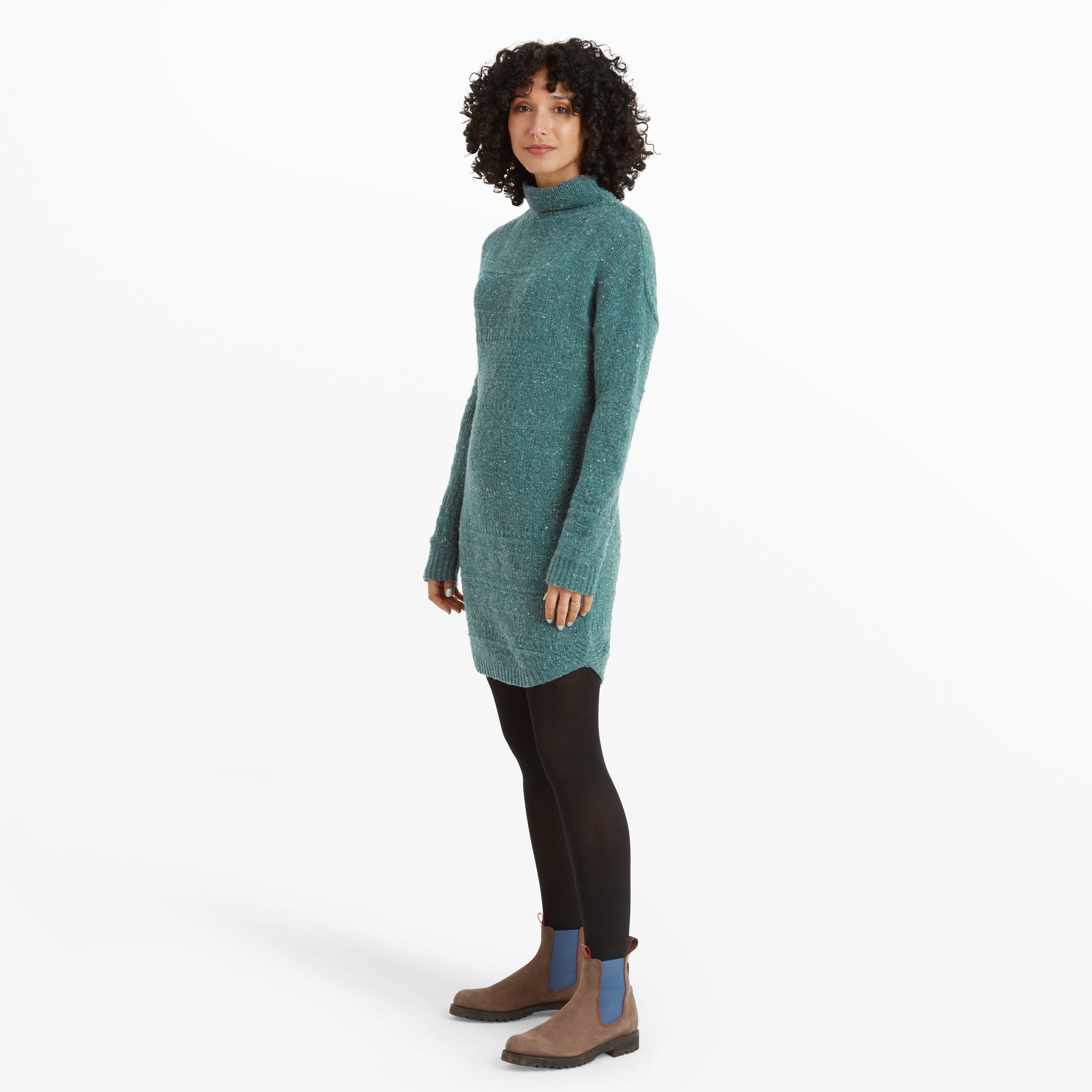 The same woman stands in profile, showcasing the side silhouette of the Sherpa Adventure Gear Yuden Dress in Blue. The hem of the dress has a subtle curve, and the turtleneck sits loosely at the neck. She pairs the outfit with the same black tights and brown Chelsea boots with blue panels.