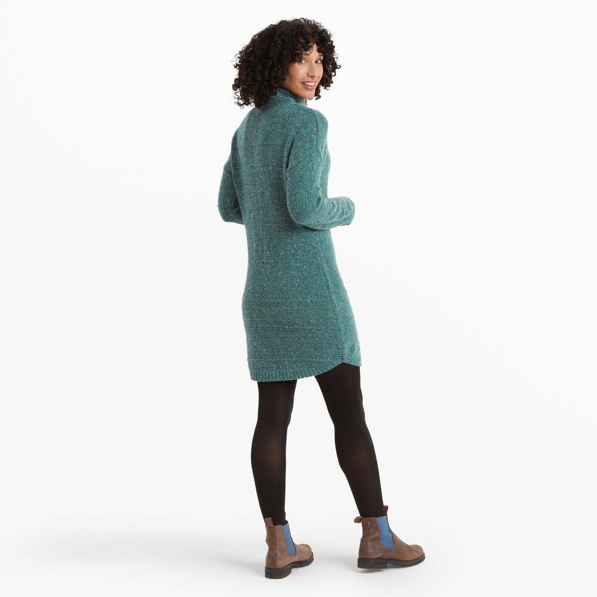 A rear view of the woman reveals the back of the Sherpa Adventure Gear Yuden Dress in Blue, emphasising its straight fit and minimalistic design. Her curly hair falls naturally, and the hemline has a slight asymmetry. She turns her head slightly back and smiles.