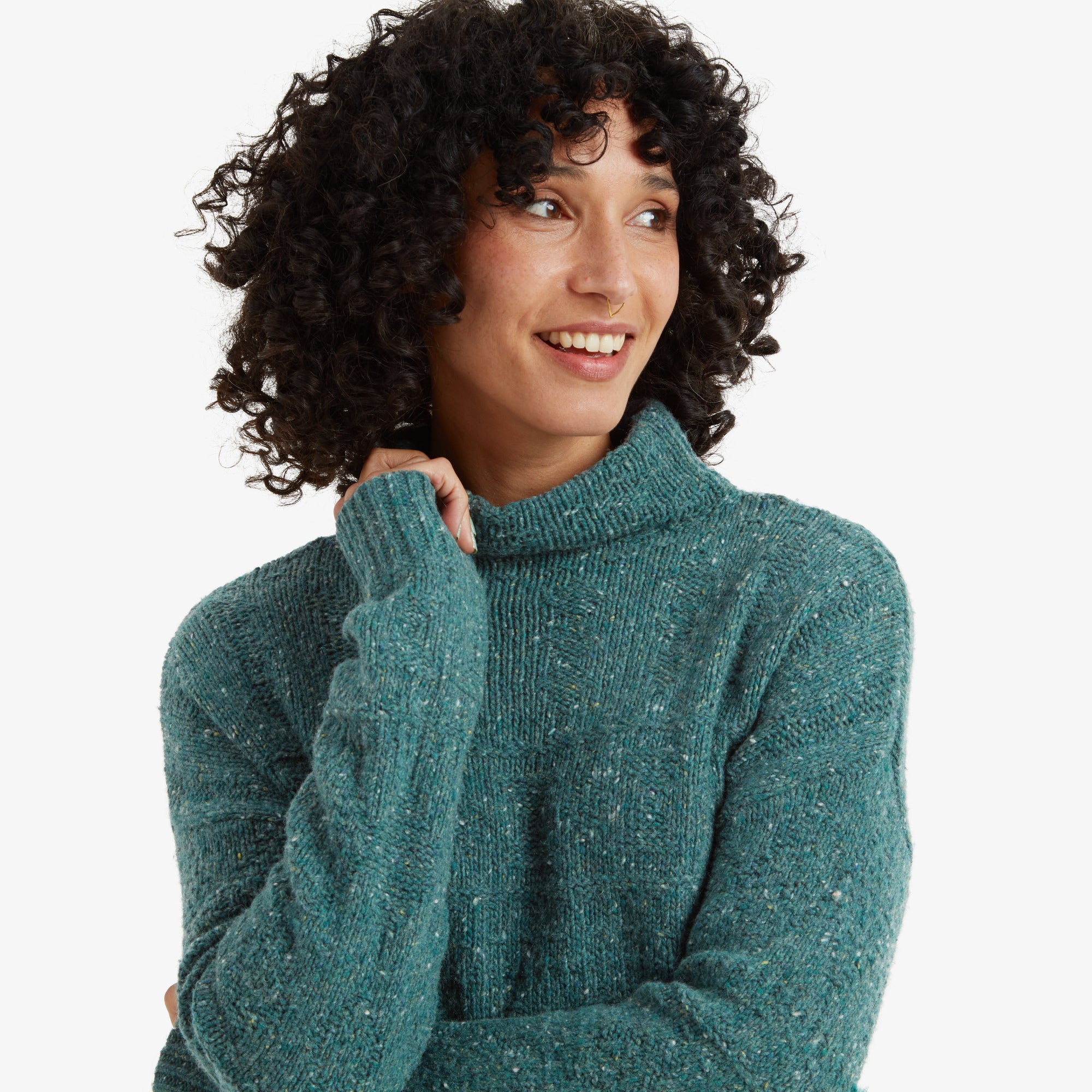 A close-up of the woman focuses on her face and the turtleneck of the Sherpa Adventure Gear Yuden Dress in Blue. The teal knit fabric shows a textured speckled pattern, and she lightly holds her arm, smiling warmly.