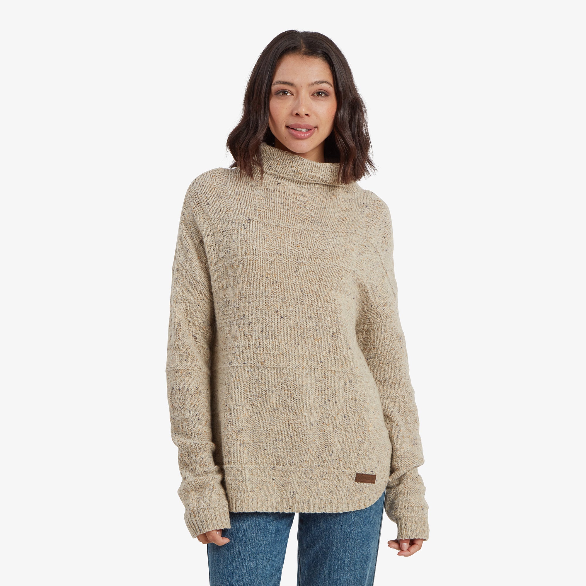 A woman is wearing a Sherpa Adventure Gear Yuden Pullover Sweater in Beige with a textured knit pattern and a loose turtleneck design. The sweater has a relaxed fit, long sleeves, and is styled with blue jeans. She is standing against a plain white background, smiling softly at the camera.