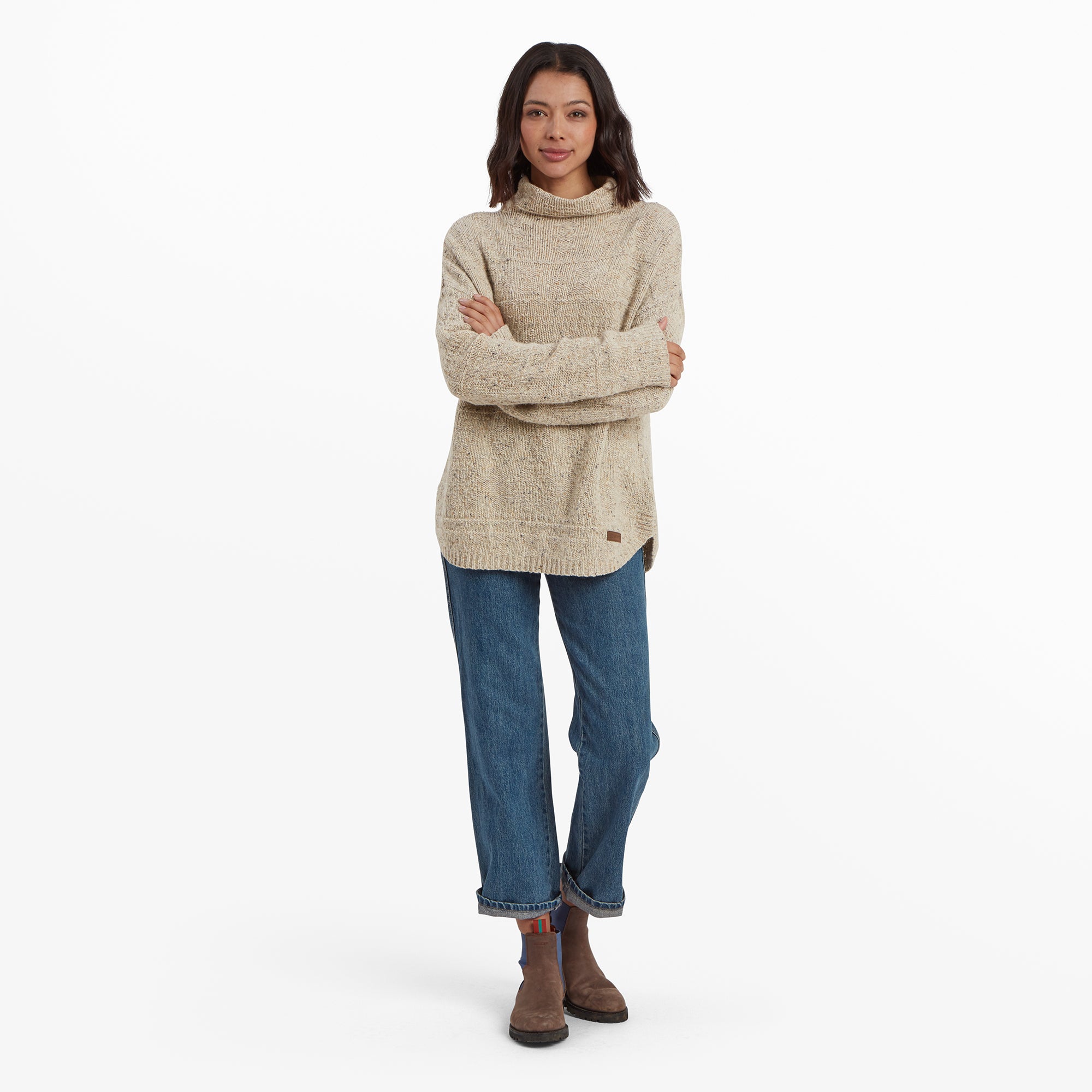 The same woman is posing in a Sherpa Adventure Gear Yuden Pullover Sweater in Beige paired with blue cuffed jeans and brown boots. She has her arms crossed, showcasing the casual and cozy fit of the sweater. The background is clean and white, emphasising the neutral tones of her outfit.