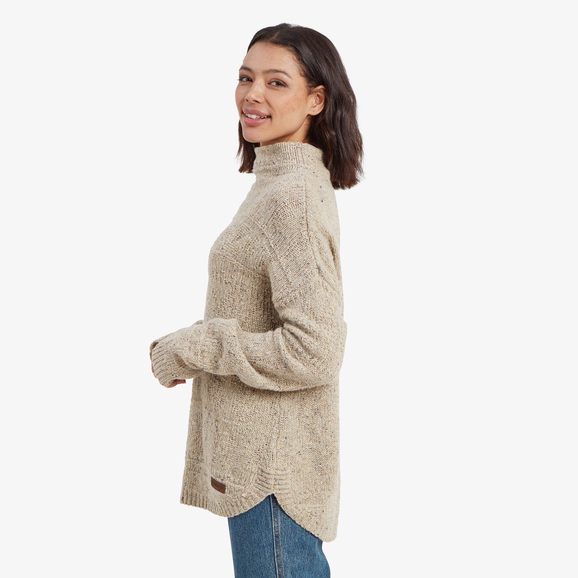 A side profile of the woman wearing the Sherpa Adventure Gear Yuden Pullover Sweater in Beige, revealing the high-low hem design with slight scalloped edges. The long sleeves drape over her hands, highlighting the sweater’s oversized, comfortable fit.