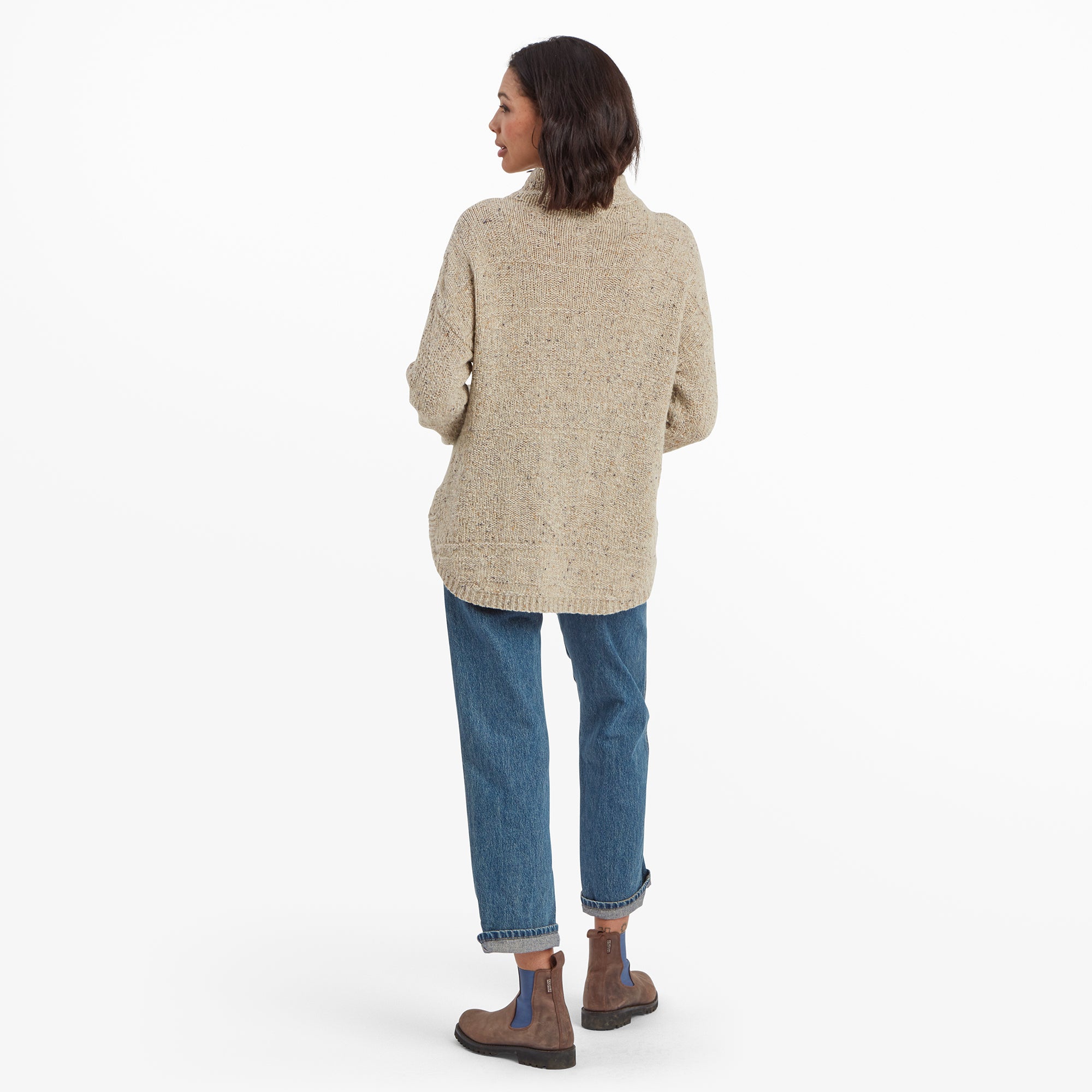 A back view of the woman modeling the Sherpa Adventure Gear Yuden Pullover Sweater in Beige, showing its full-length coverage and simple knit pattern. The jeans and boots complement the laid-back aesthetic of the sweater.