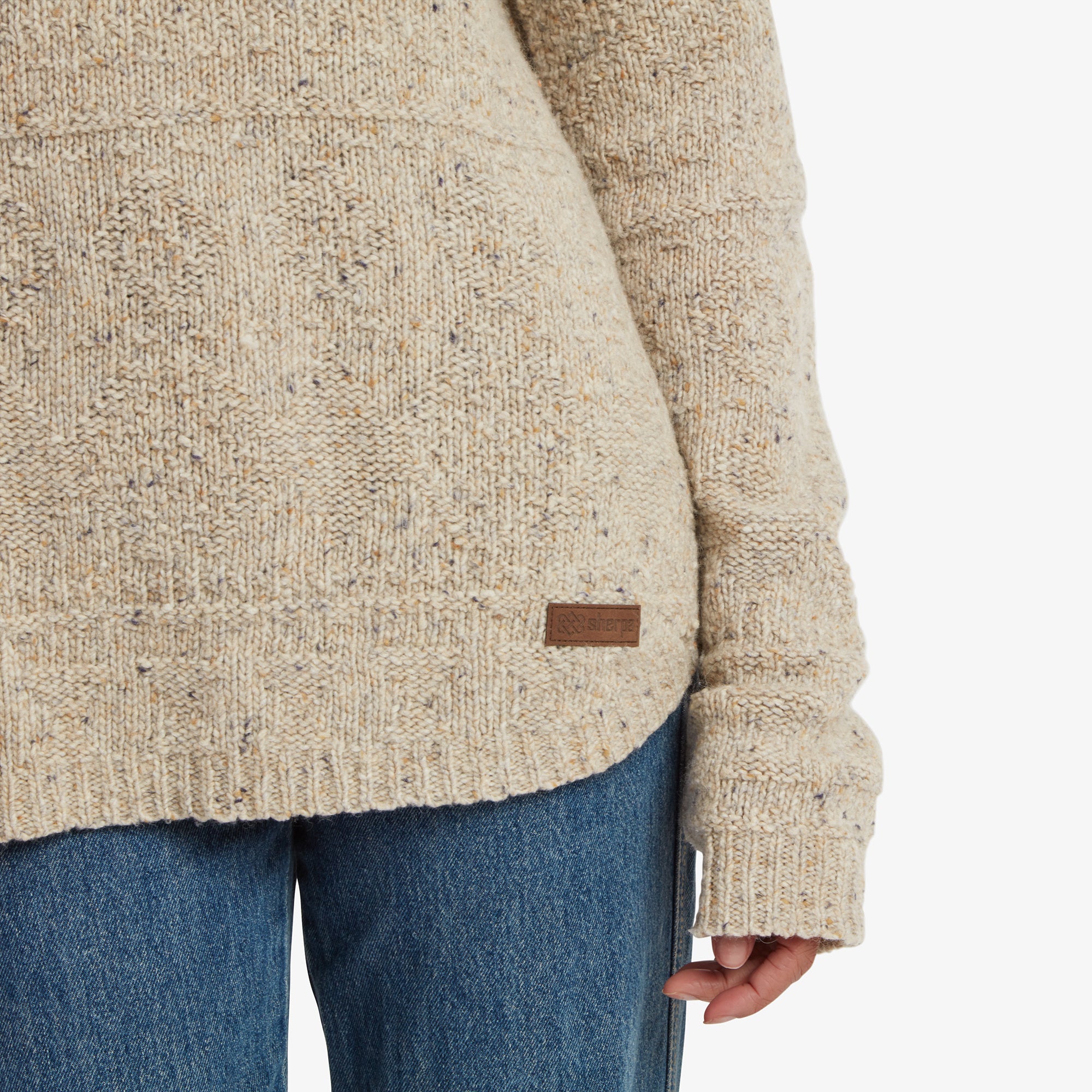 A close-up of the lower portion of the Sherpa Adventure Gear Yuden Pullover Sweater in Beige, highlighting the ribbed hem and subtle brand logo on a brown leather patch. The jeans complete the cozy and casual look.