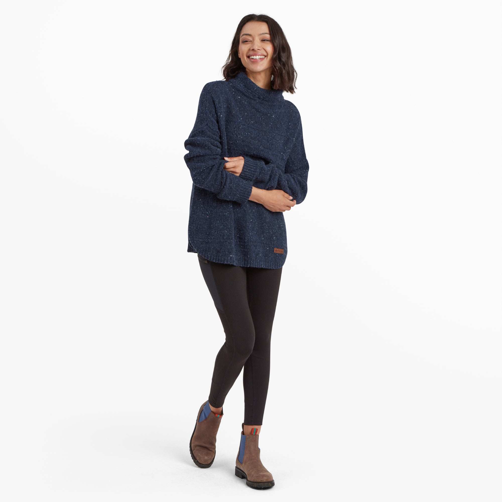 A full-body view of the model in a Sherpa Adventure Gear Yuden Pullover Sweater in Blue and black leggings. The model is standing in a relaxed pose with arms folded and wearing brown ankle boots with blue accents, creating a casual yet stylish look.