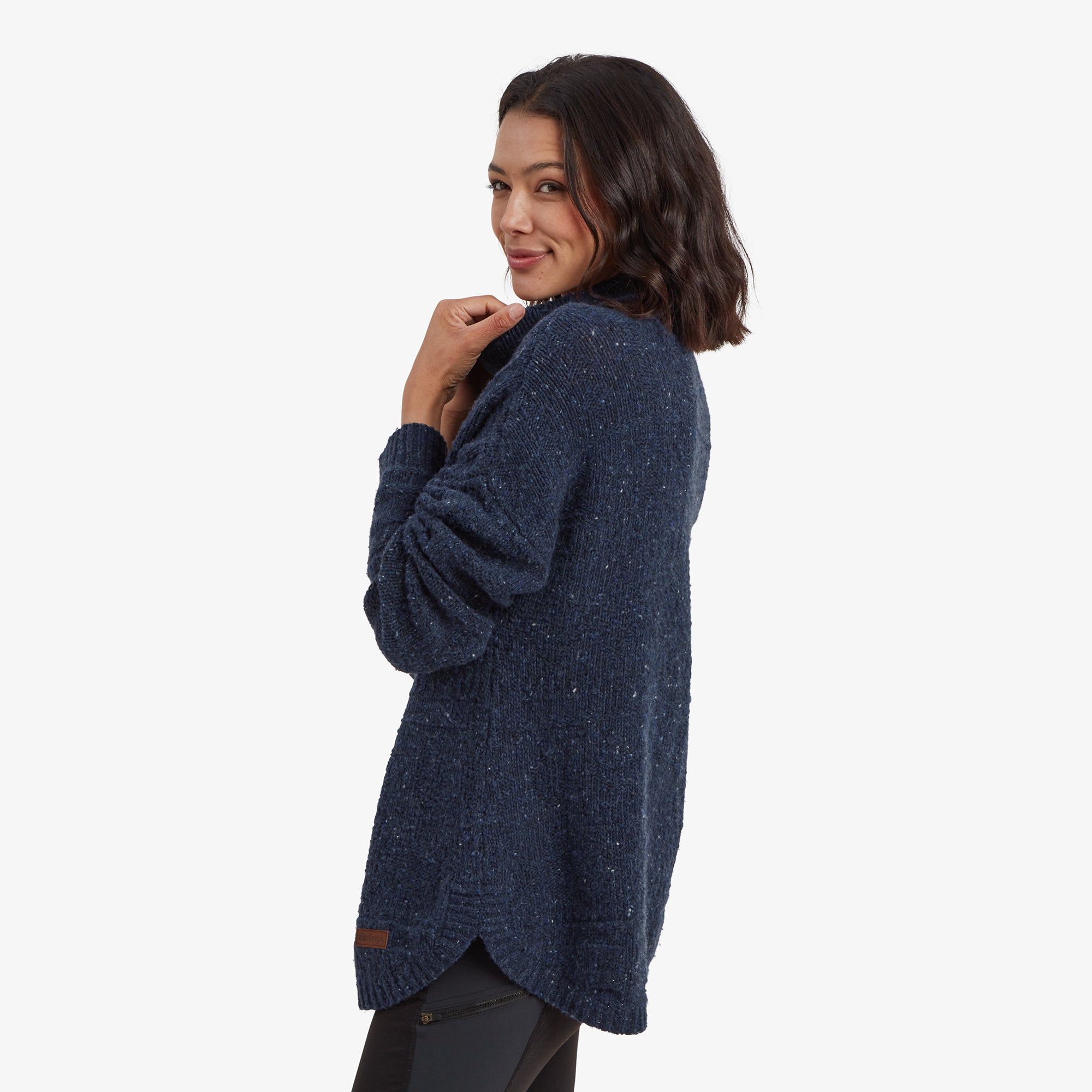 A side view of the model in the Sherpa Adventure Gear Yuden Pullover Sweater in Blue, highlighting the curved hemline that dips slightly longer in the back. The texture of the sweater is visible, showing intricate knitting patterns and white flecks in the fabric.